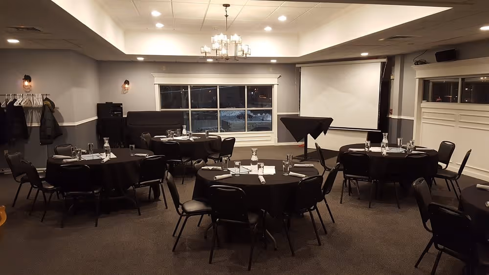 Banquet room with round tables covered in black tablecloths, each set with glasses and napkins, under warm ceiling lights.
