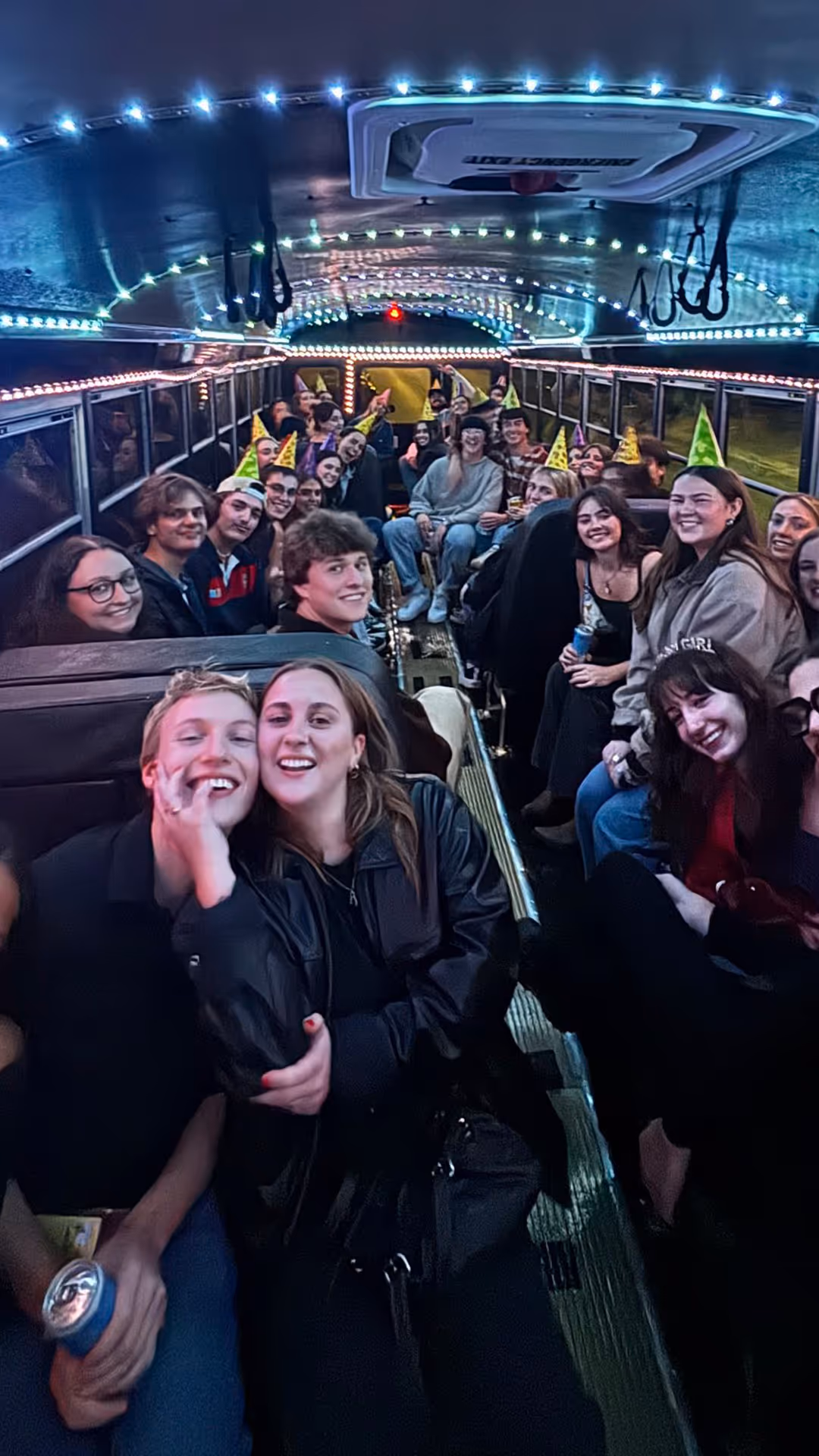 Group of young people wearing party hats, smiling, and sitting inside a party bus decorated with dynamic nightclub lights.