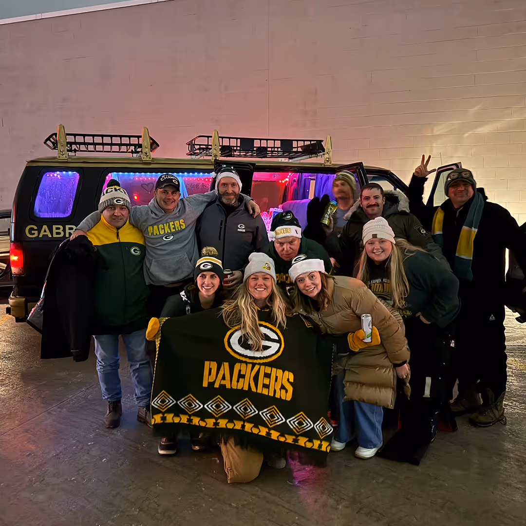 Group of people dressed in Green Bay Packers gear posing in front of a party van with colorful lights inside, holding a Packers blanket and drinks.
