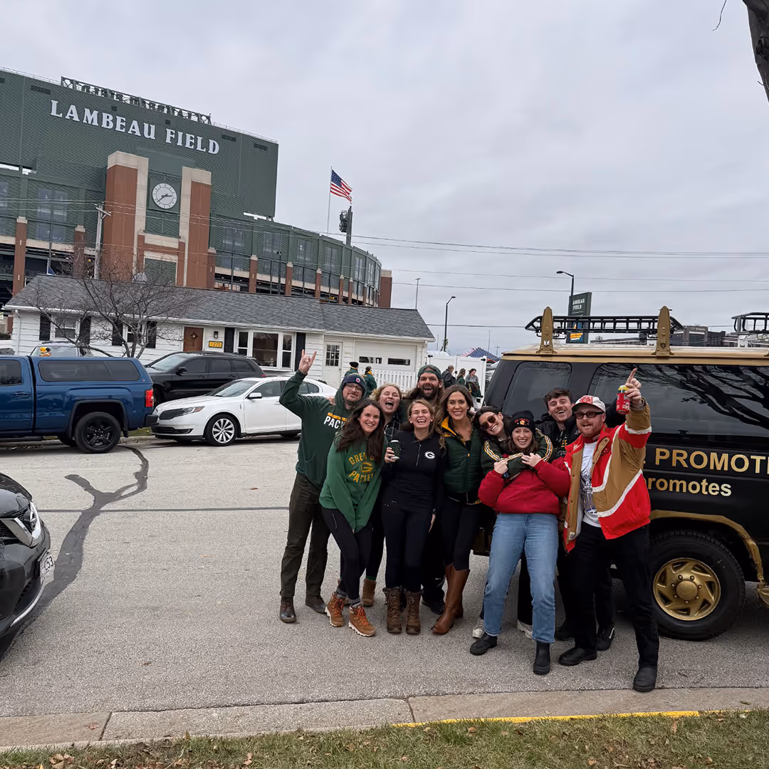 Group of nine happy people posing together in front of Lambeau Field stadium and a party van on a cloudy day.