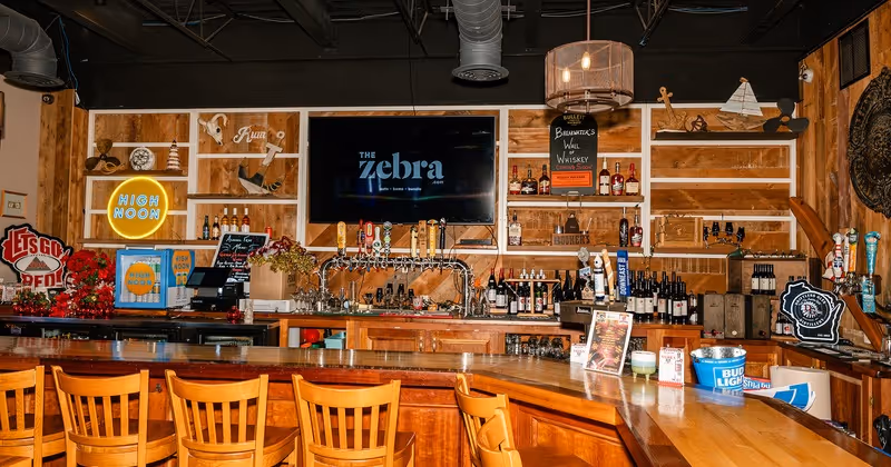 Interior view of a wooden bar with chairs, multiple liquor bottles, beer taps, a TV displaying 'The zebra' logo, and various decorative signs including a neon 'High Noon' sign.