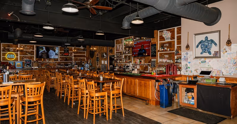 Interior of a cozy bar with wooden tables and chairs, a long wooden bar counter, shelves with bottles, and TV screens on the walls.