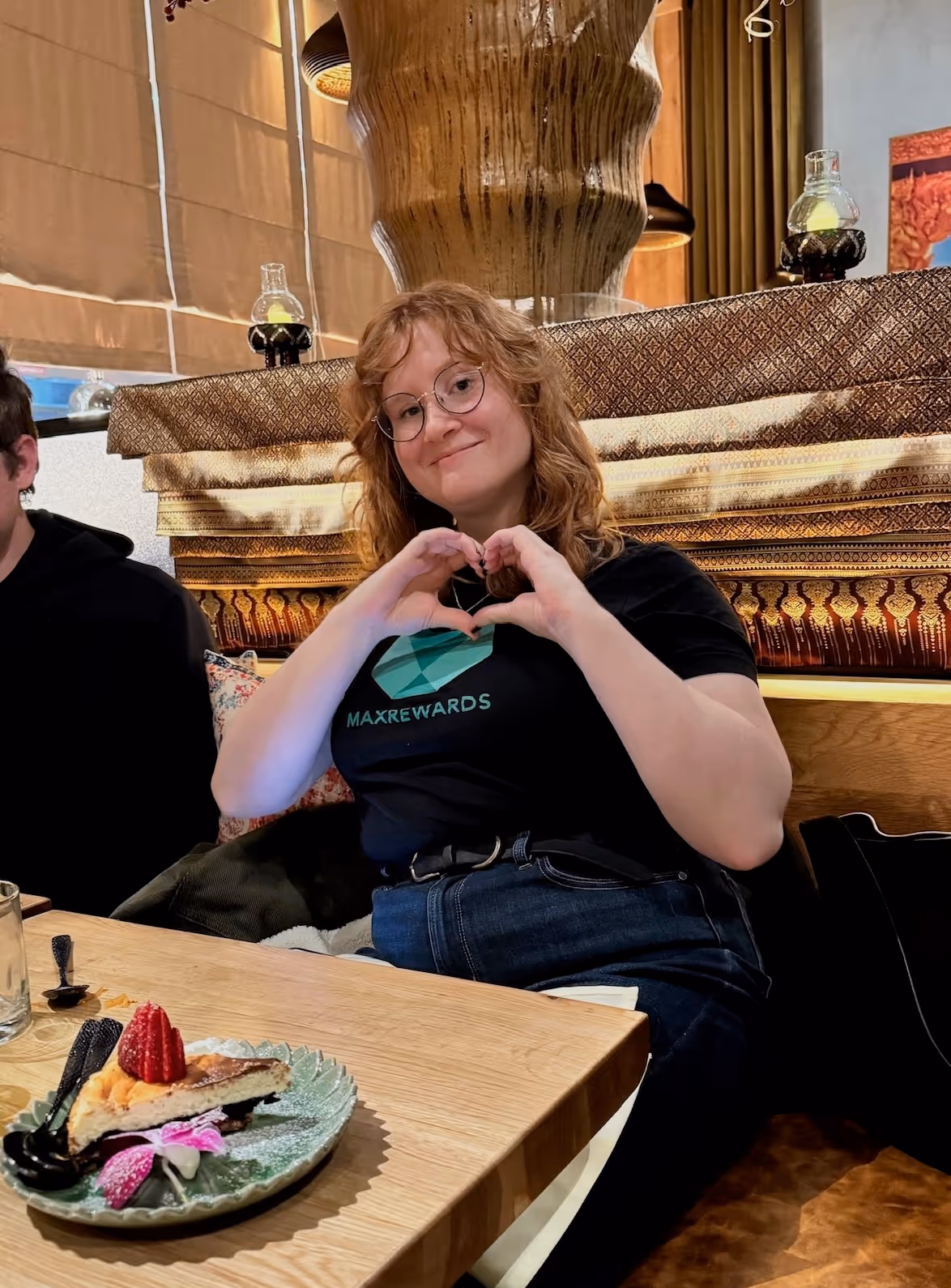 Smiling woman with glasses wearing a MaxRewards t-shirt, sitting at a table making a heart shape with her hands in a cozy restaurant setting.