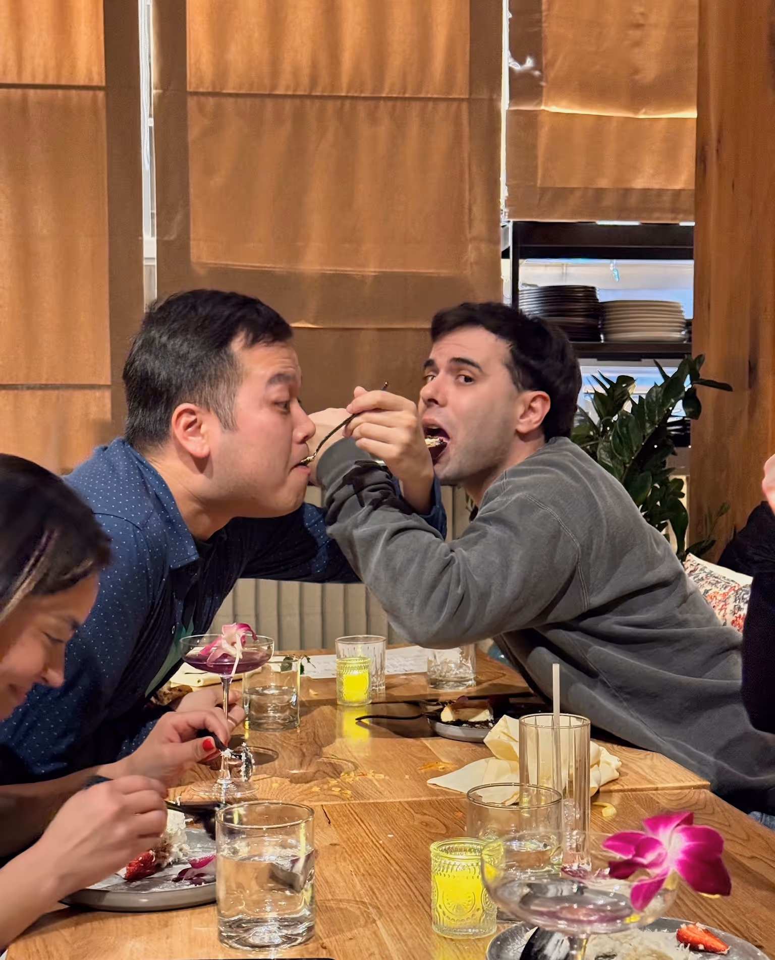 Two men playfully feeding each other with spoons while sitting at a wooden dining table with glasses and plates of food.