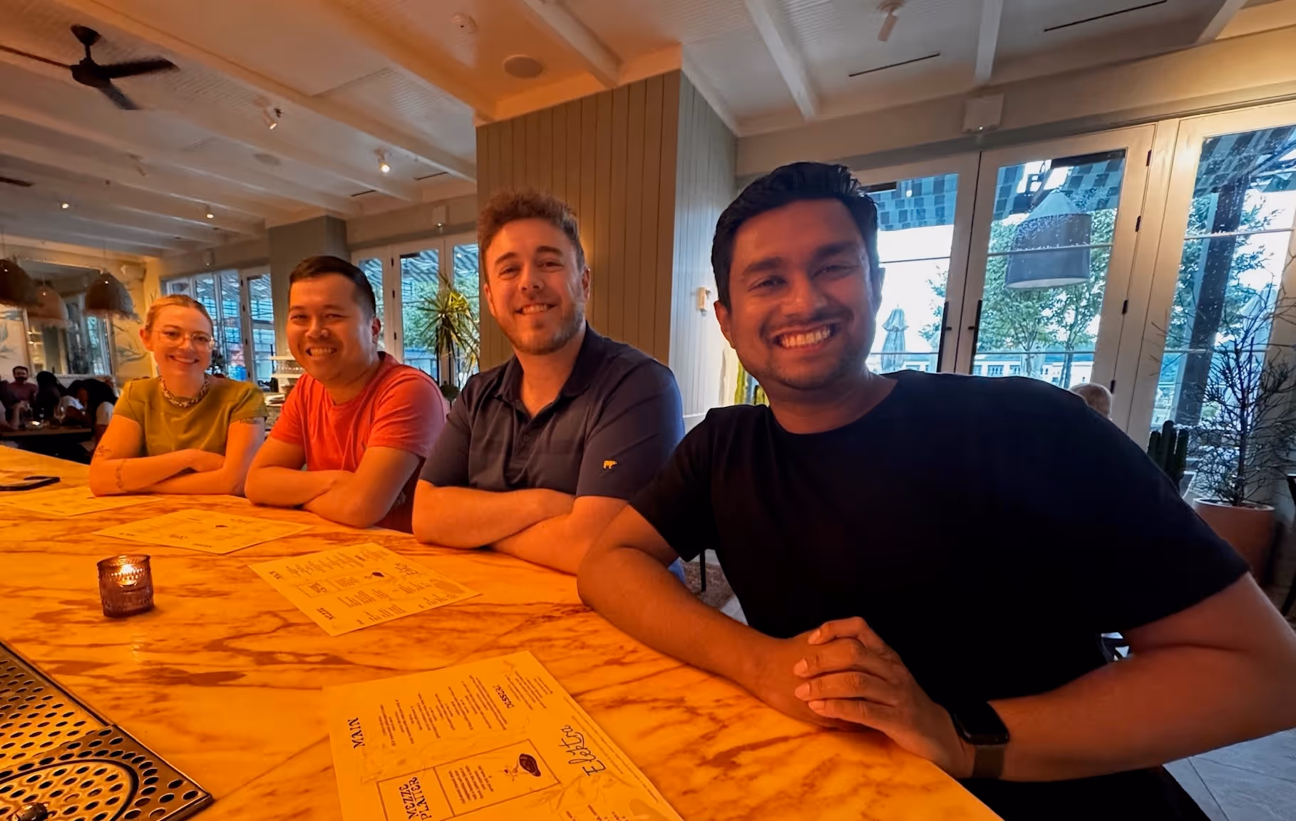 Four people smiling and sitting side by side at a marble countertop with menus in a warmly lit restaurant.