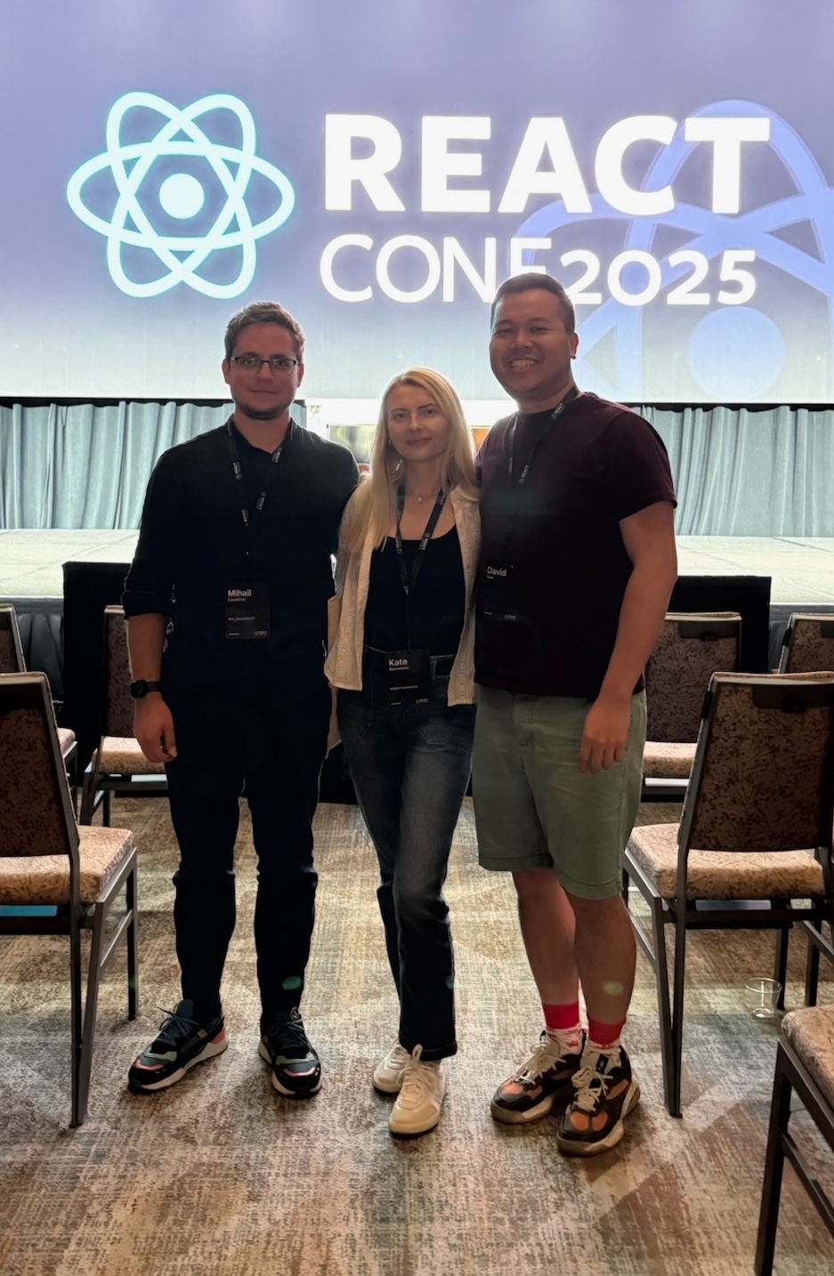 Three conference attendees standing and smiling in front of a stage with a React Conf 2025 logo.