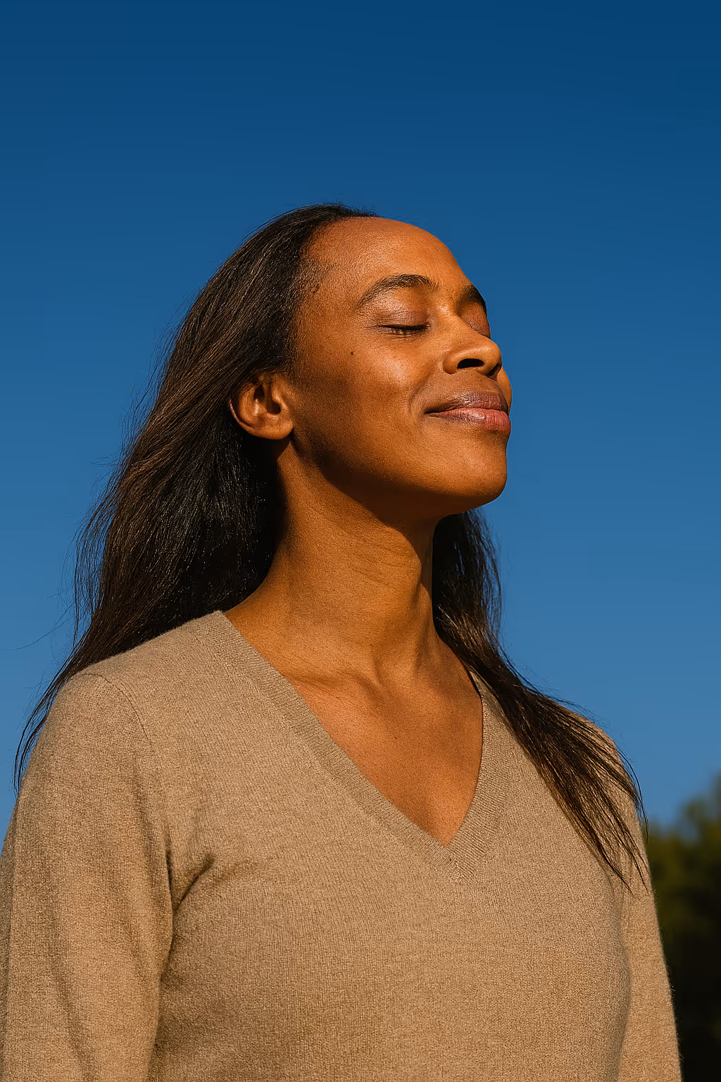 Woman with long hair and a beige sweater standing outdoors with eyes closed and a serene expression against a clear blue sky.