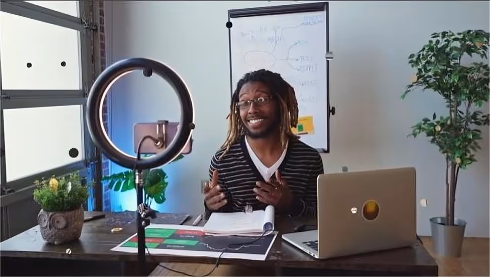 Man with glasses and dreadlocks speaking in front of a laptop and ring light in a home office setting.