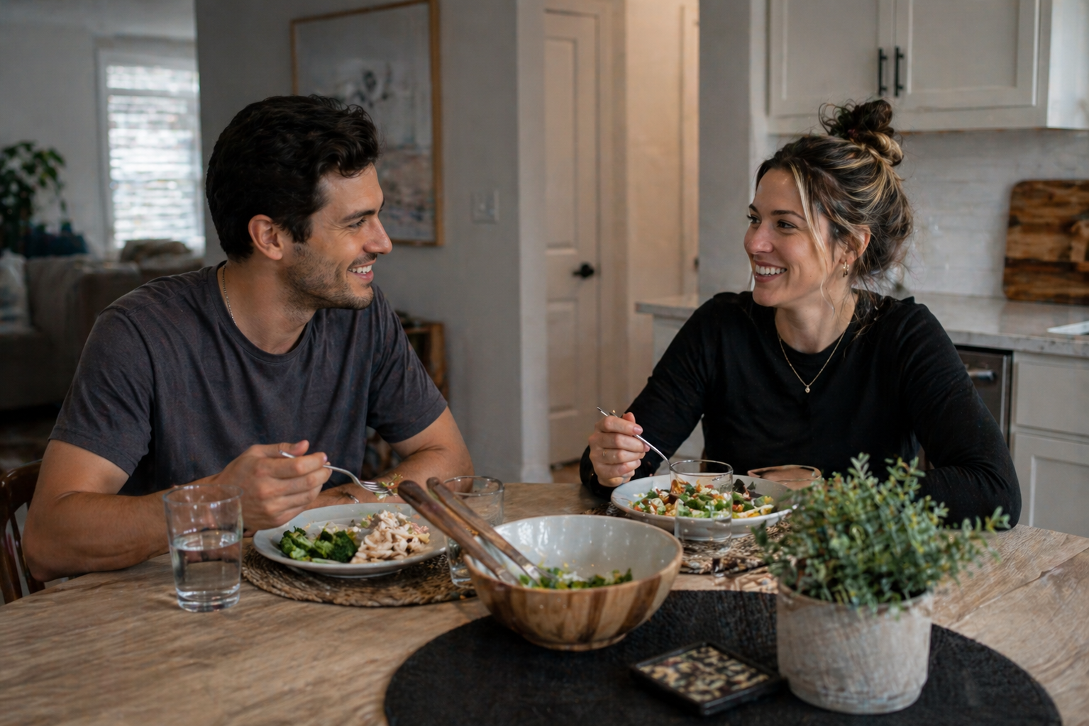 Two people in recovery sitting at a kitchen table together in a sober living home, sharing a meal and laughing, representing peer community and daily structure in addiction recovery