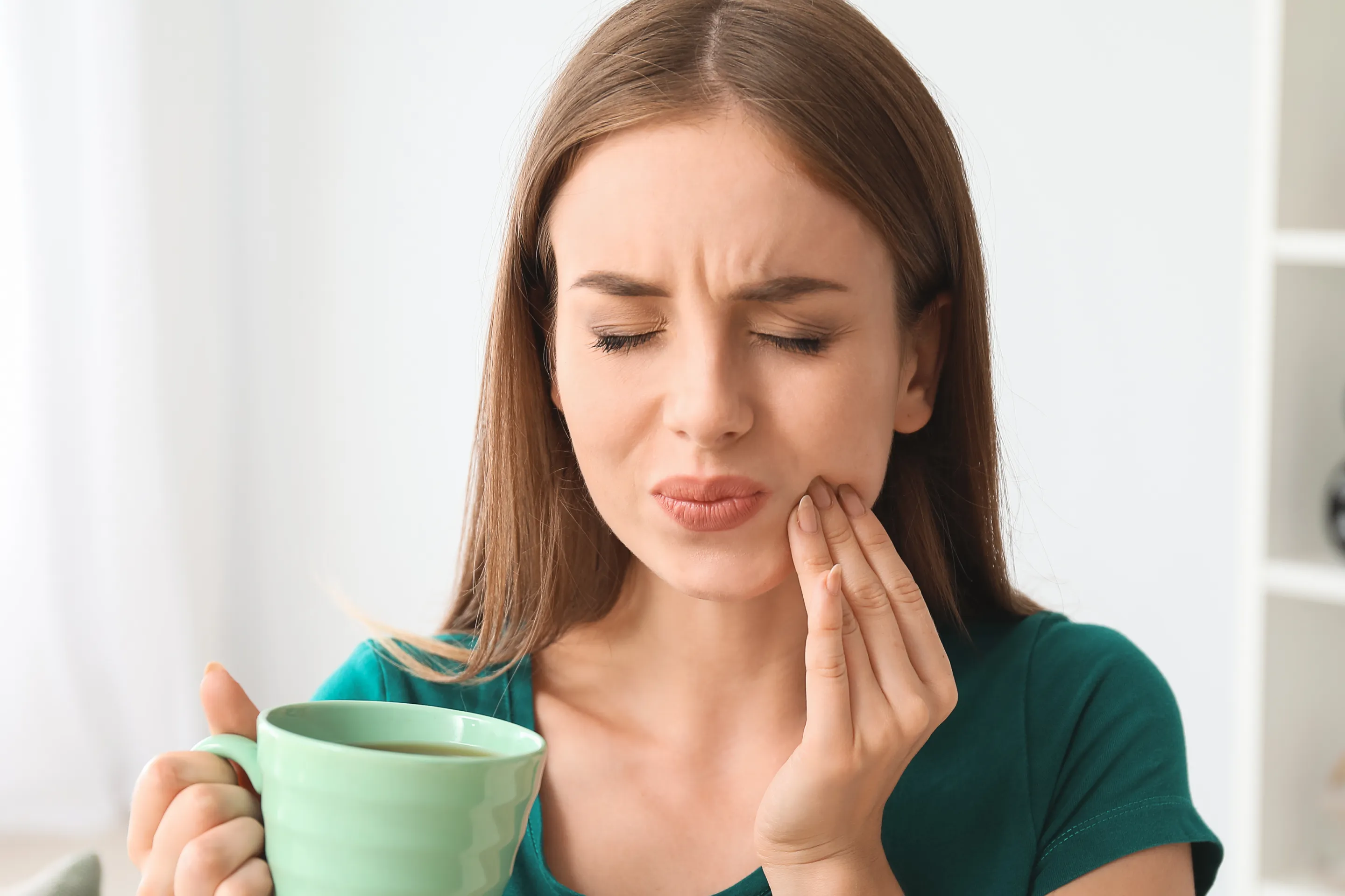 A woman holds her cheek in pain while holding a green mug in the other hand.