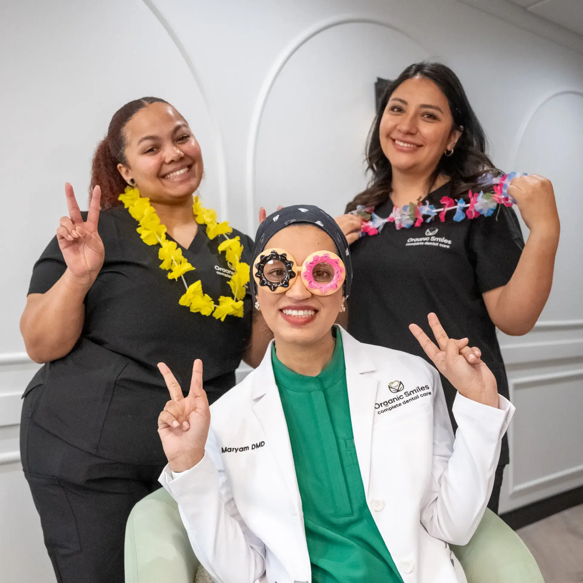 Three women smile and pose with peace signs, wearing colorful leis and playful eye covers.