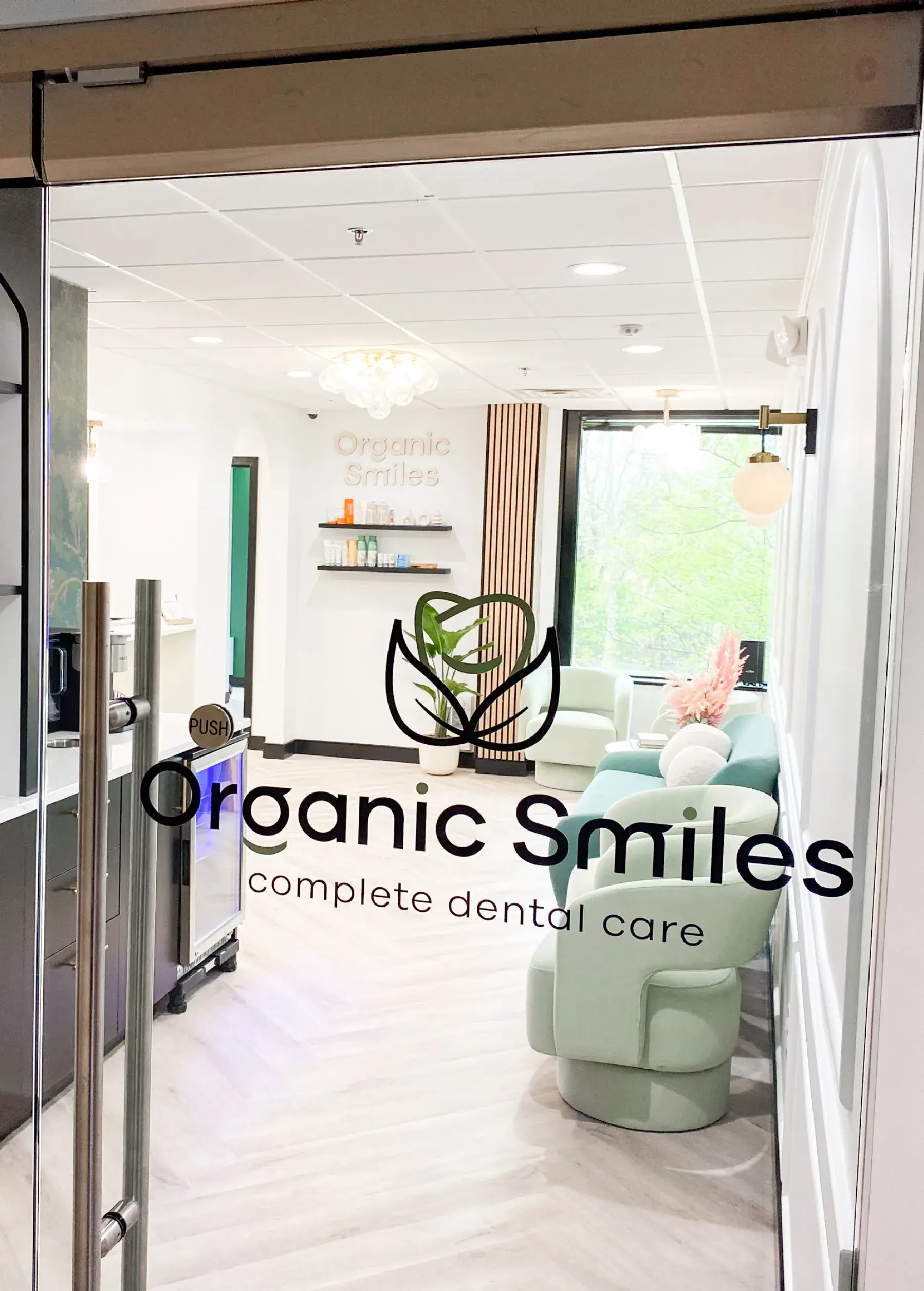 Dental office entrance with glass doors and modern interior, featuring logo "Organic Smiles" and chairs inside.