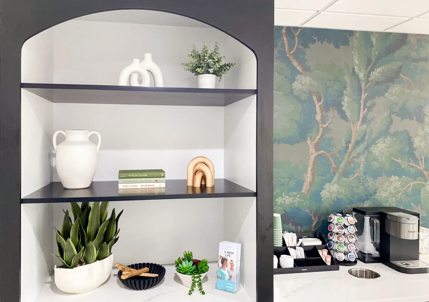 A modern shelf with plants, books, and ceramic objects next to a coffee station with supplies.