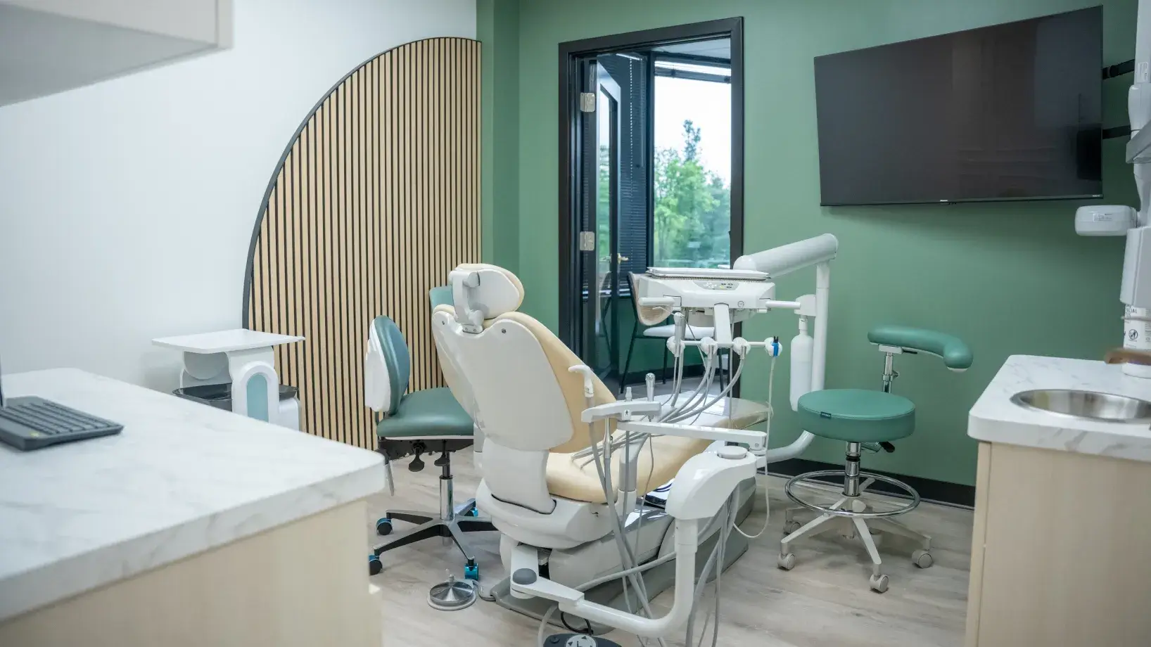 Dental office with a beige chair, dental tools, and a green wall with a mounted screen.