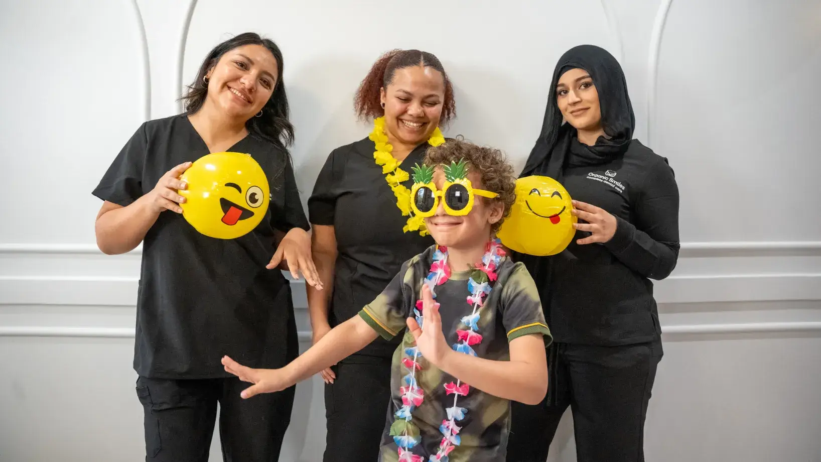 Child wearing sunglasses and lei smiles with three women holding emoji balloons, all looking happy.