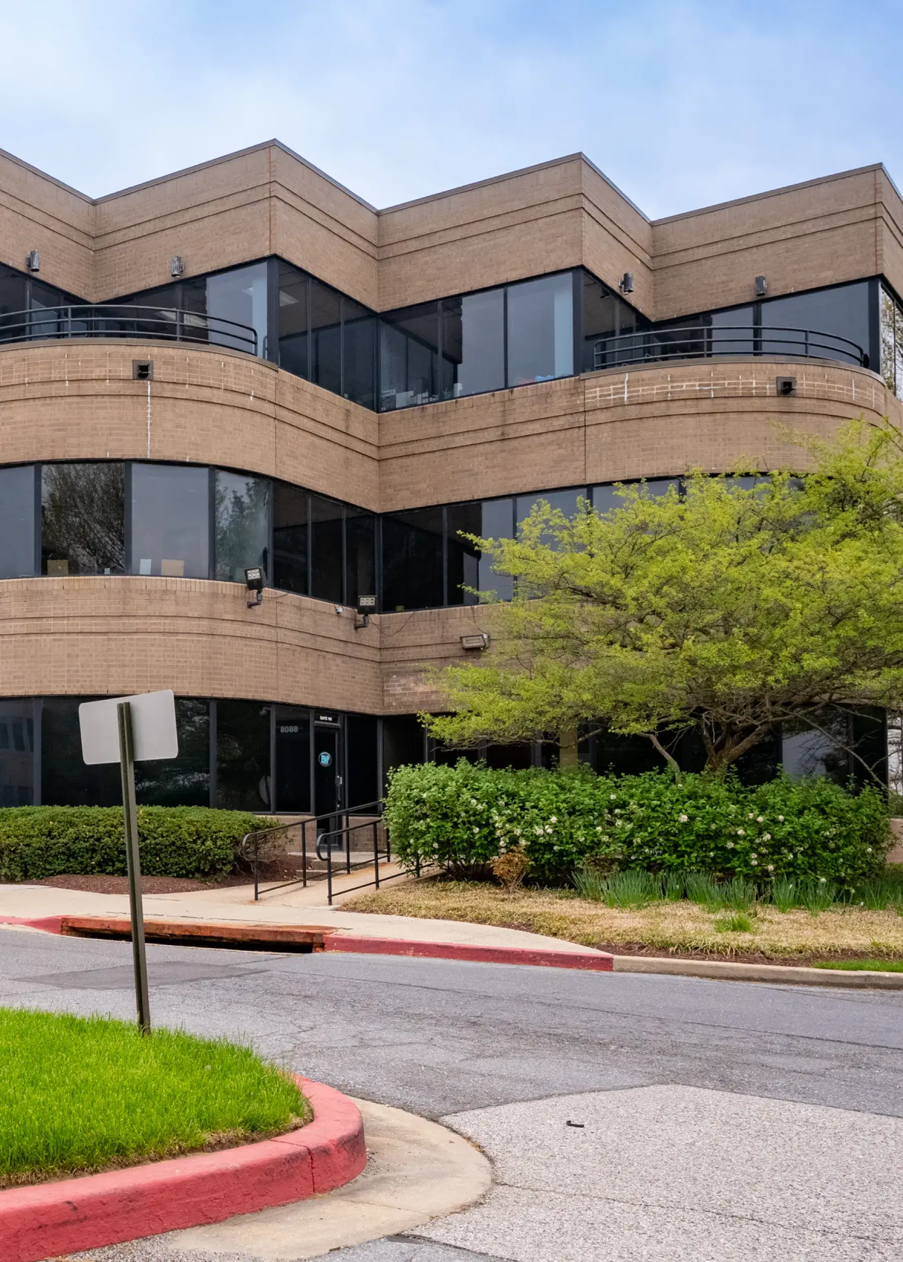 Three-story brown office building with large black windows, surrounded by greenery and a sidewalk.