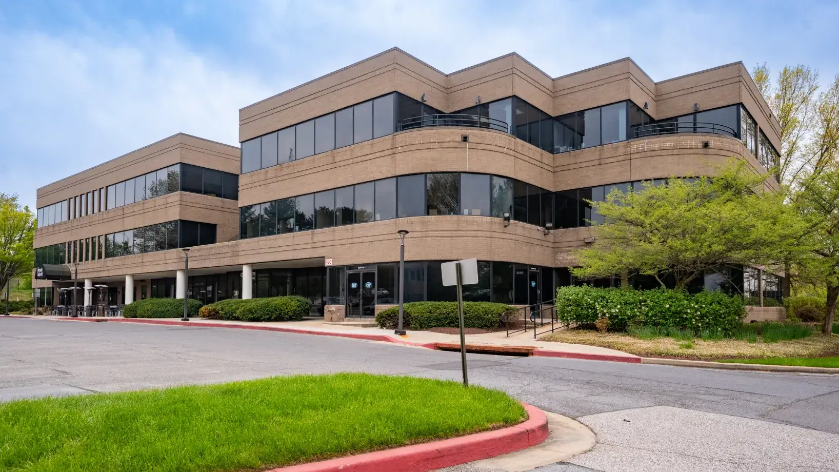 Three-story office building with large windows, surrounded by greenery and a parking lot in front.