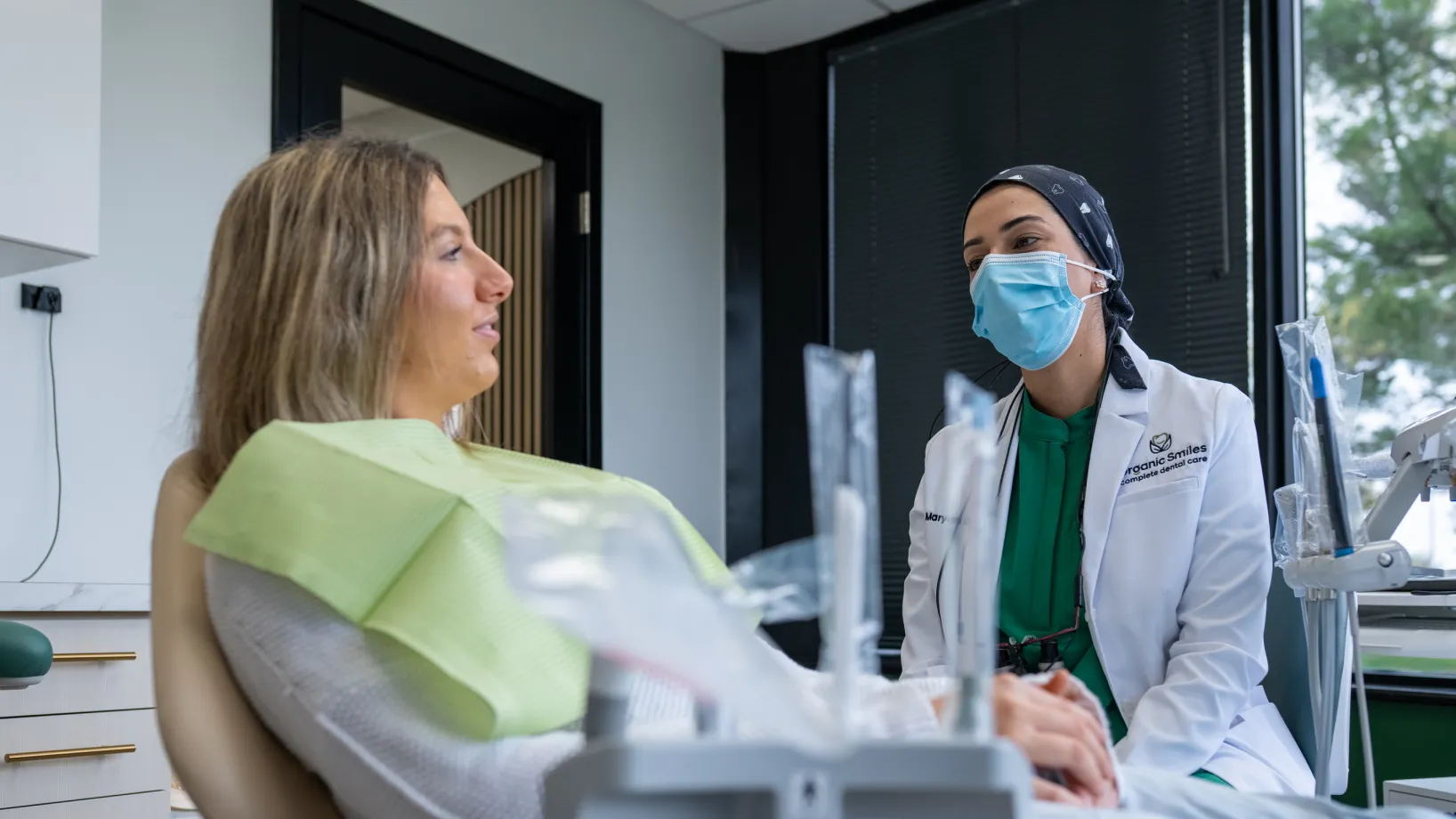 Dentist wearing a mask talks to a seated patient in a dental clinic.