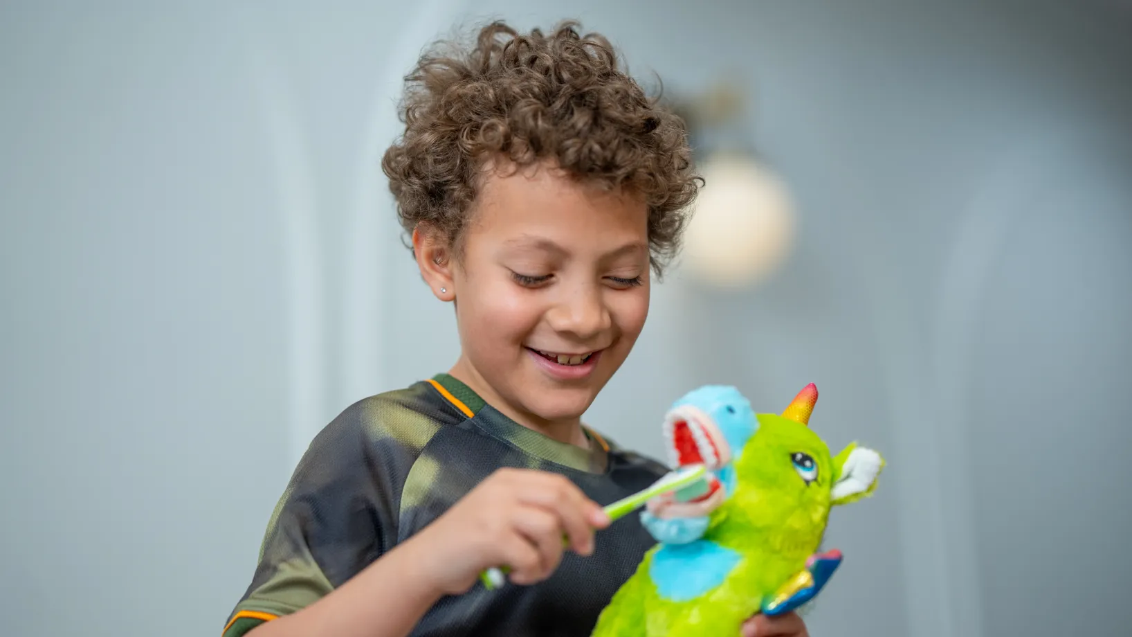 A child smiles while brushing a colorful toy dinosaur's teeth with a toothbrush.