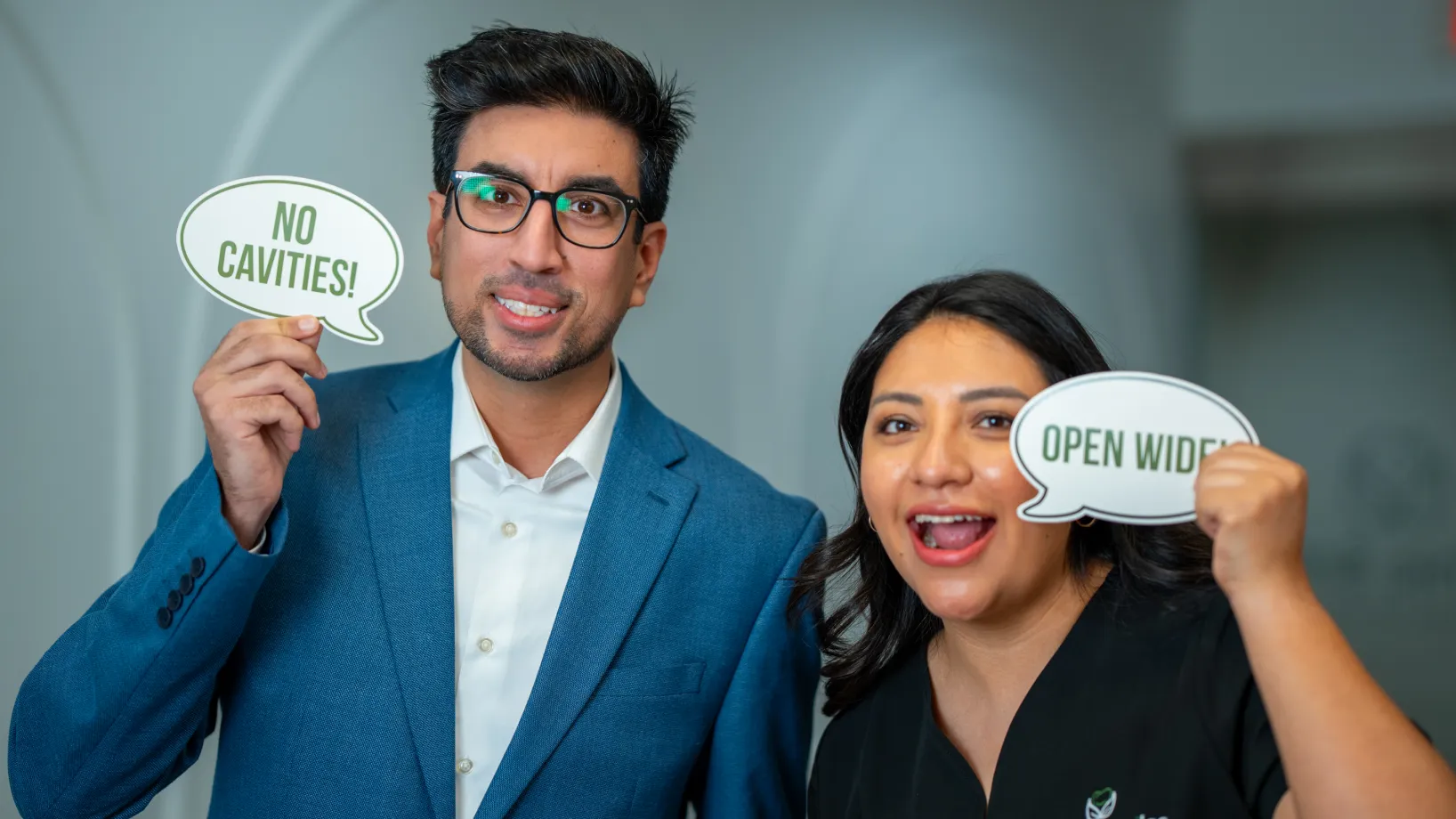 Two people smiling and holding signs that say "No Cavities!" and "Open Wide."