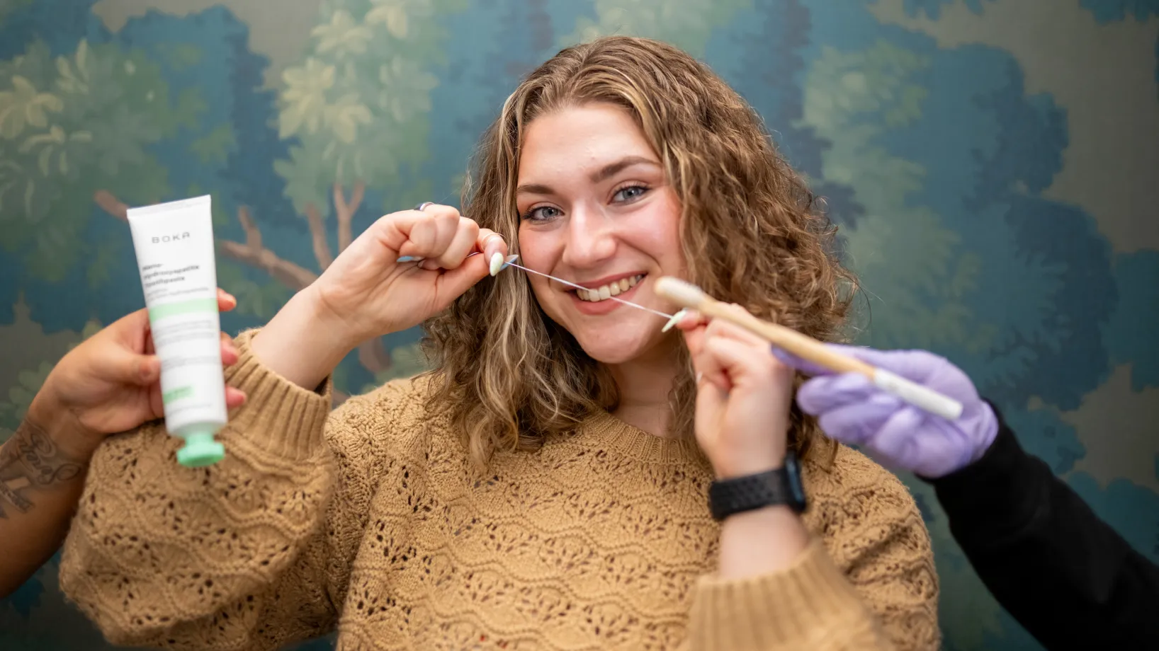 A smiling person holds dental floss with toothbrushes and toothpaste nearby.
