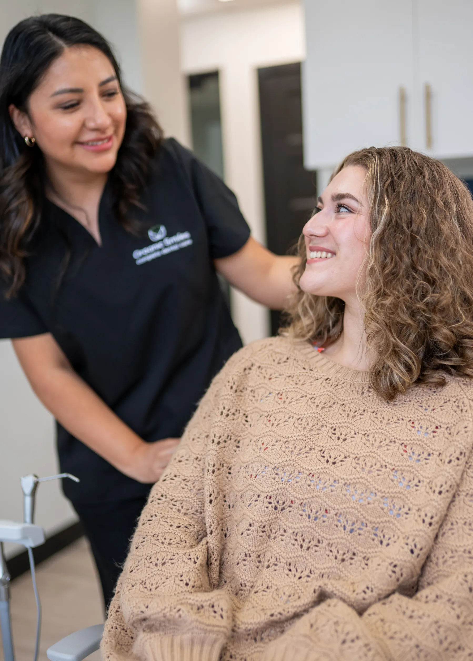 A dental professional smiles and talks to a seated patient in a dental office.