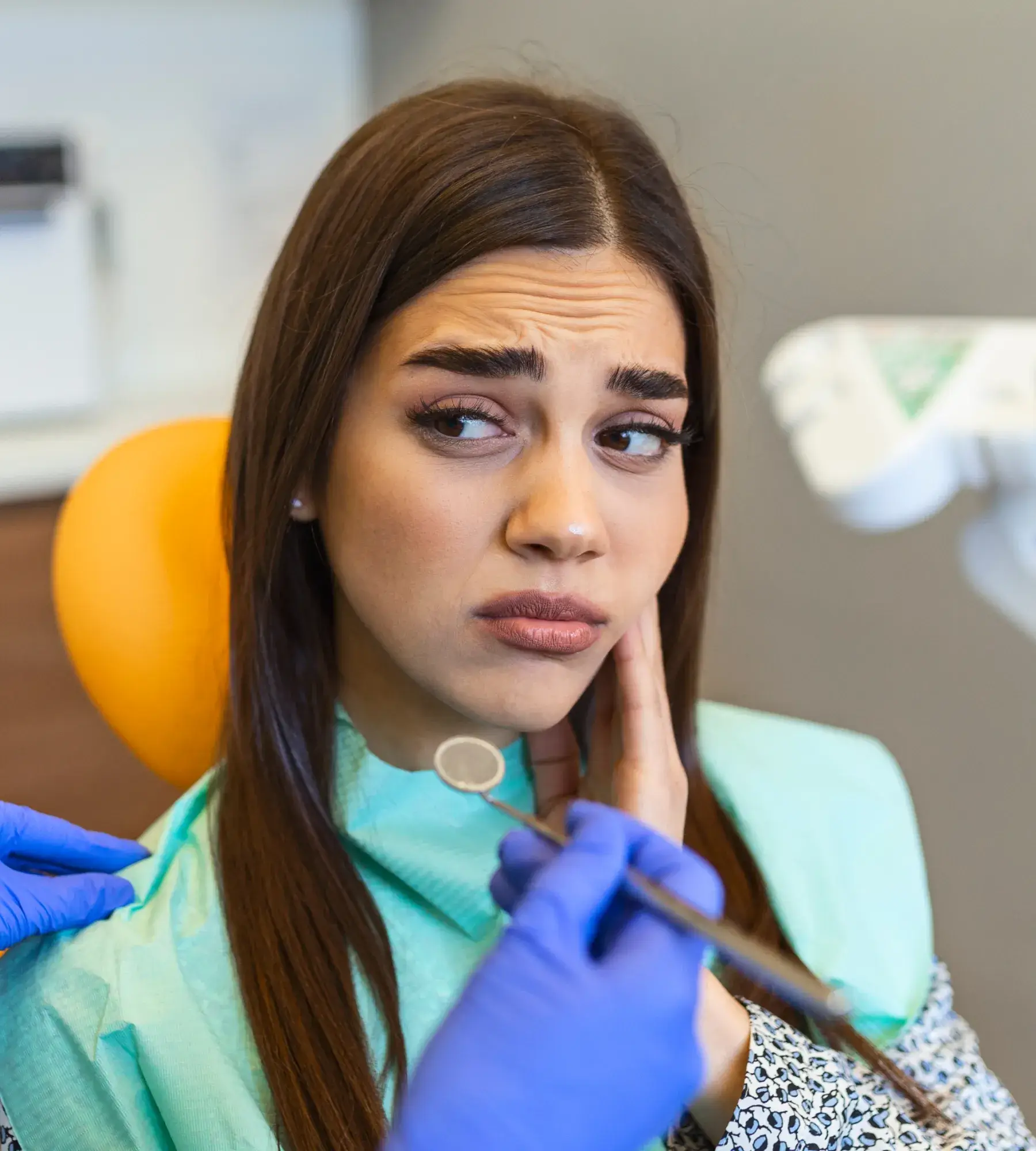 A woman looks worried, touching her face in a dental office while a dentist holds tools.
