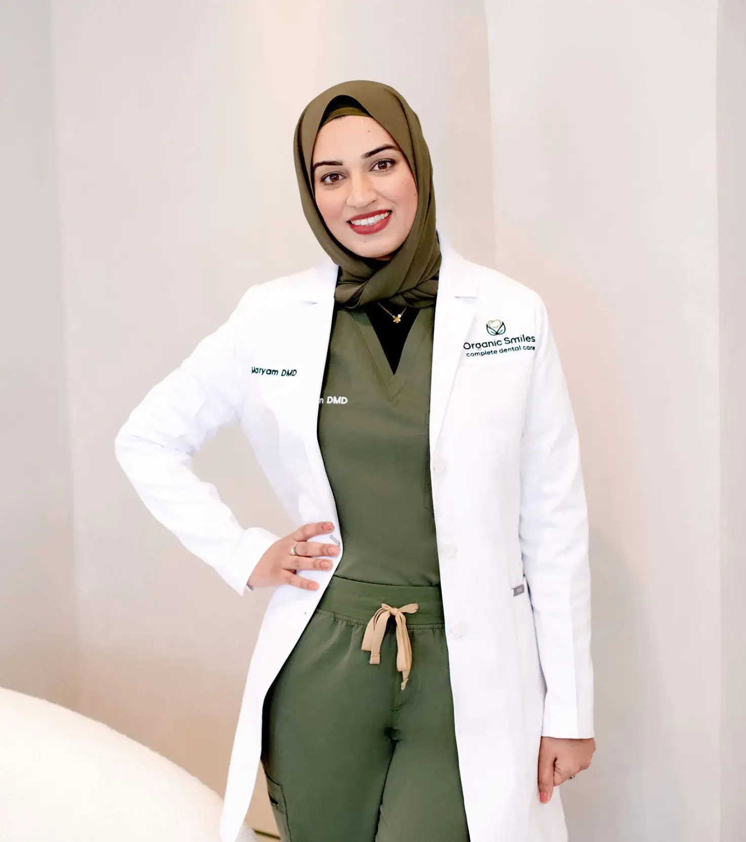 A person in a white coat and green scrubs stands smiling with a hand on their hip.