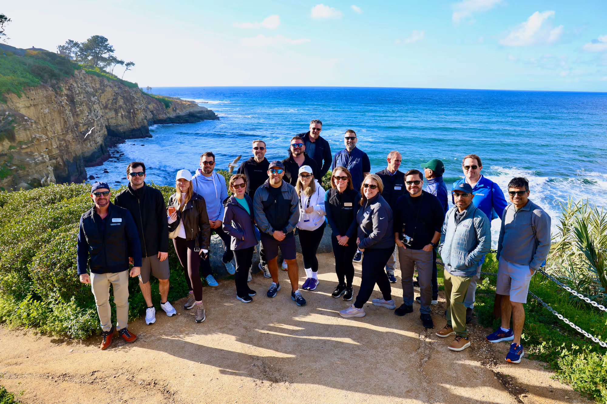 Group of 19 people posing on a coastal trail with cliffs and the ocean in the background under a partly cloudy sky.