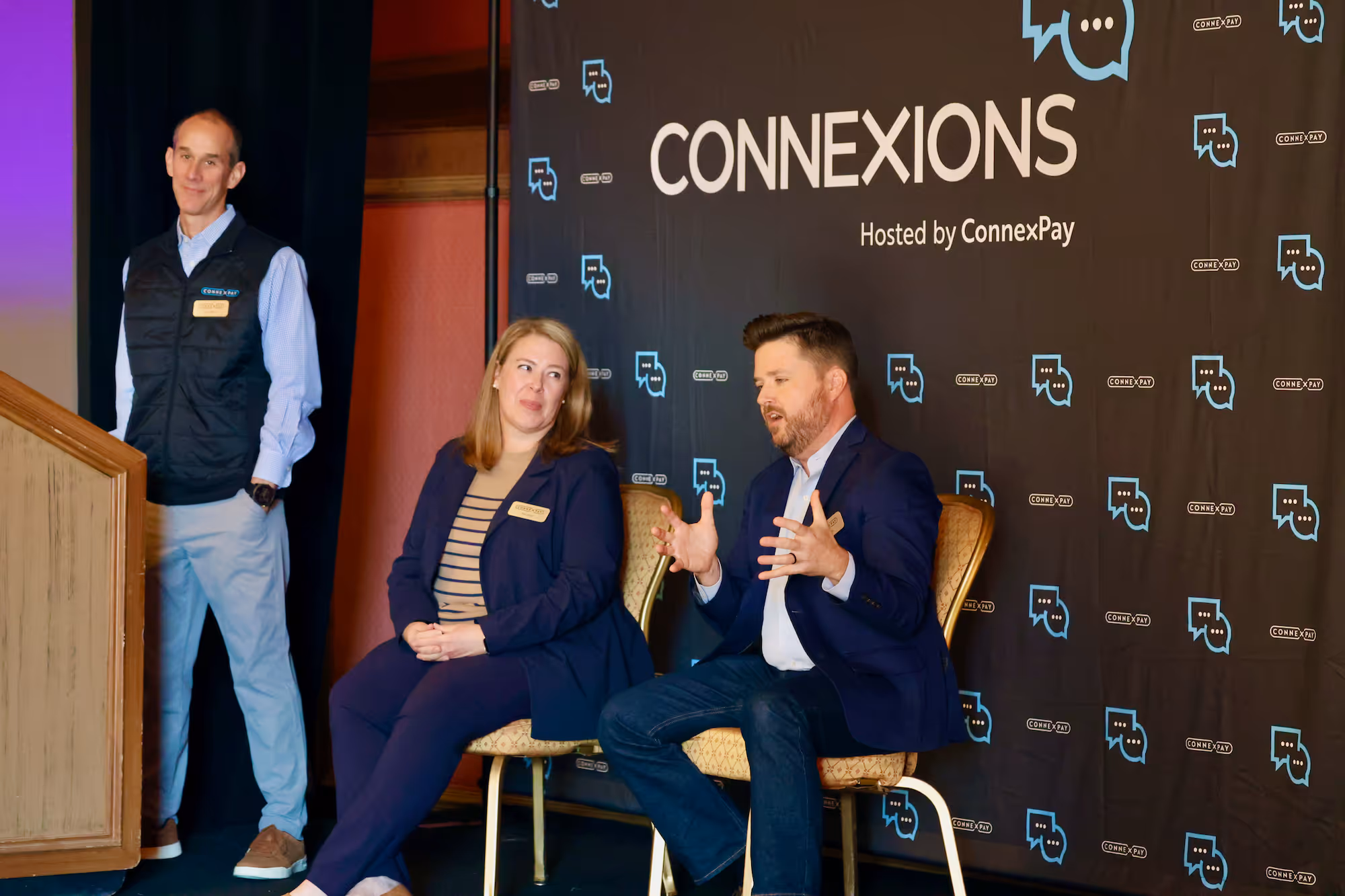 Man standing next to a podium and two people seated in chairs having a discussion at a Connexions event hosted by ConnexPay.