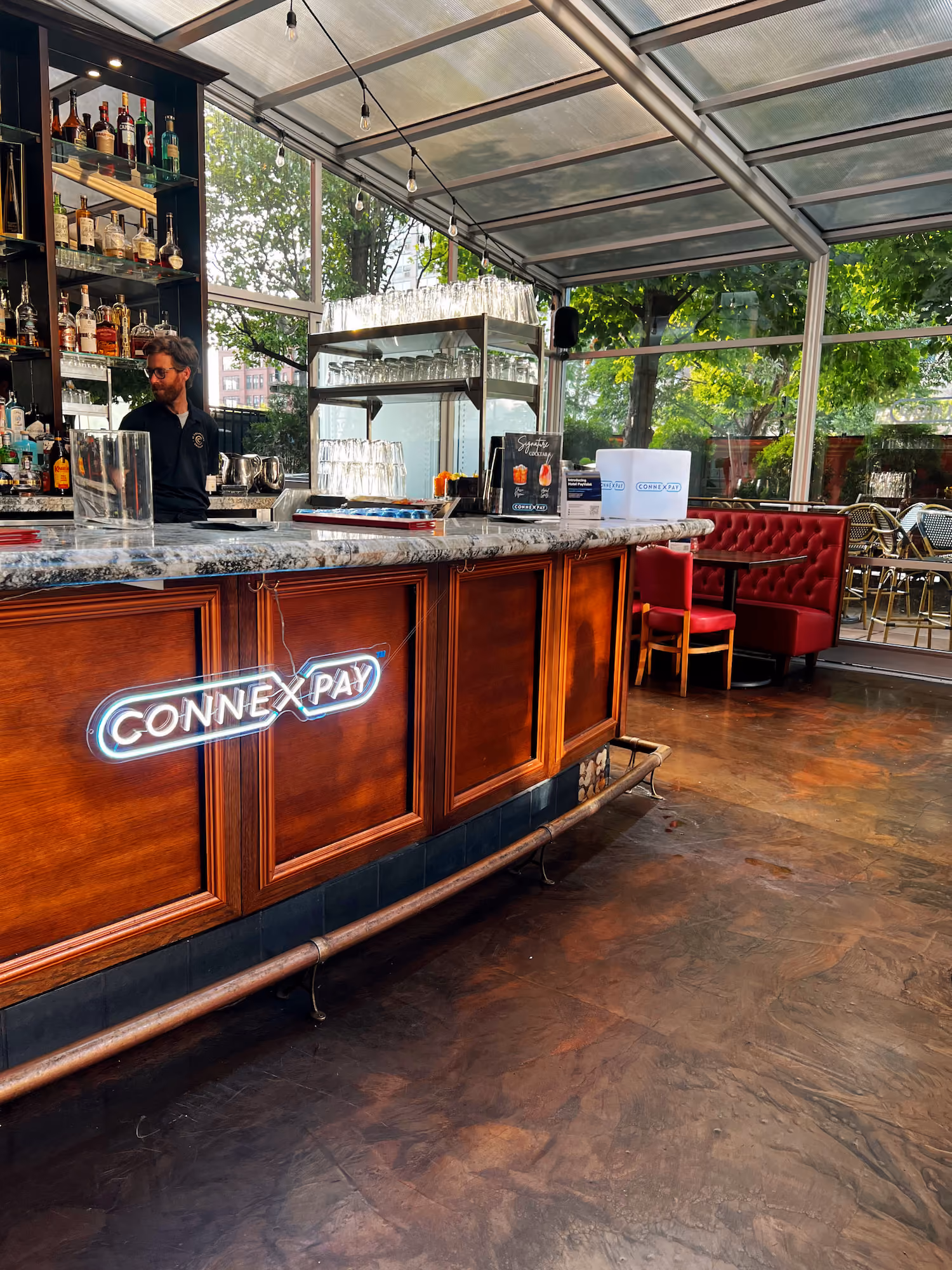 Bar counter with countertop and liquor bottles, a bartender, and red seating booths near large windows.