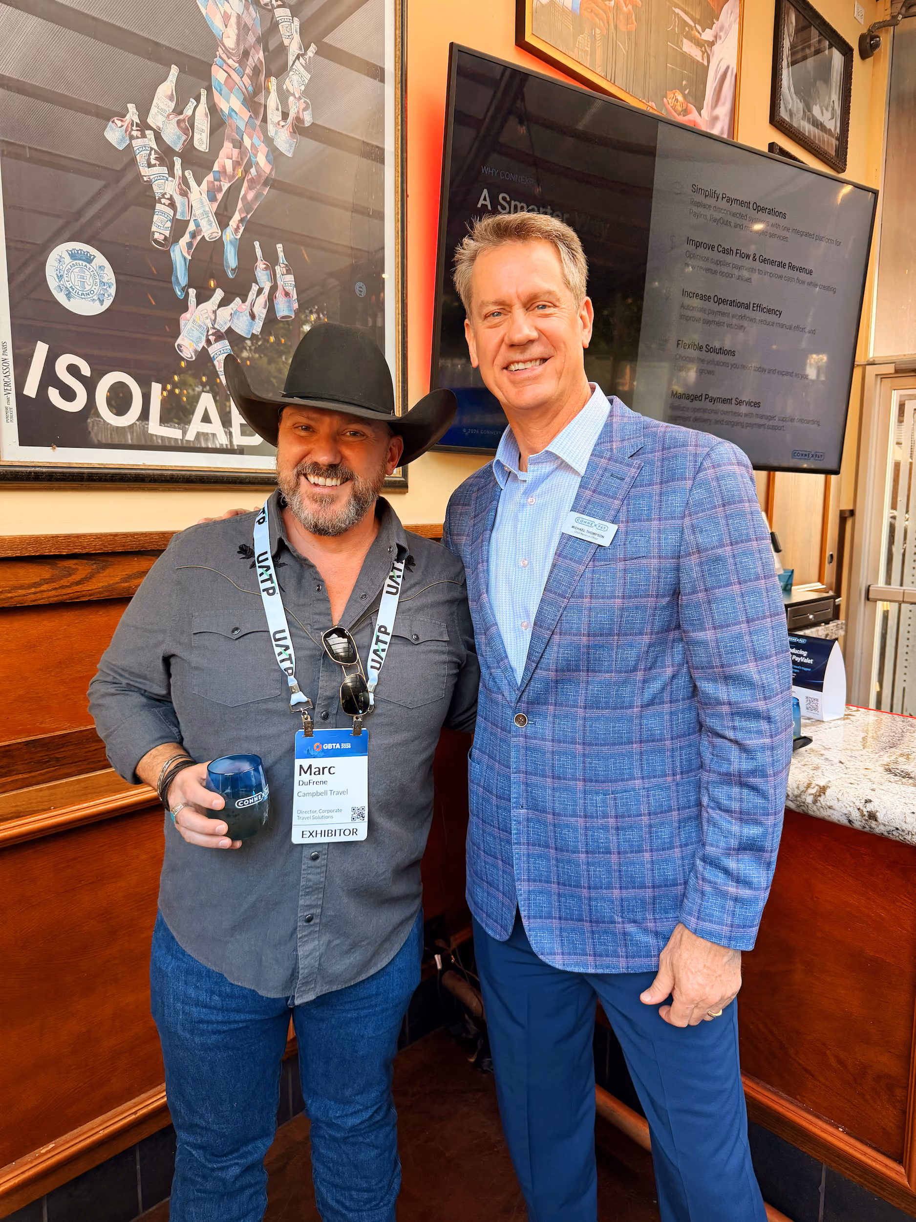 Two men smiling indoors, one in a black hat and gray shirt with an exhibitor badge, the other in a blue plaid jacket.