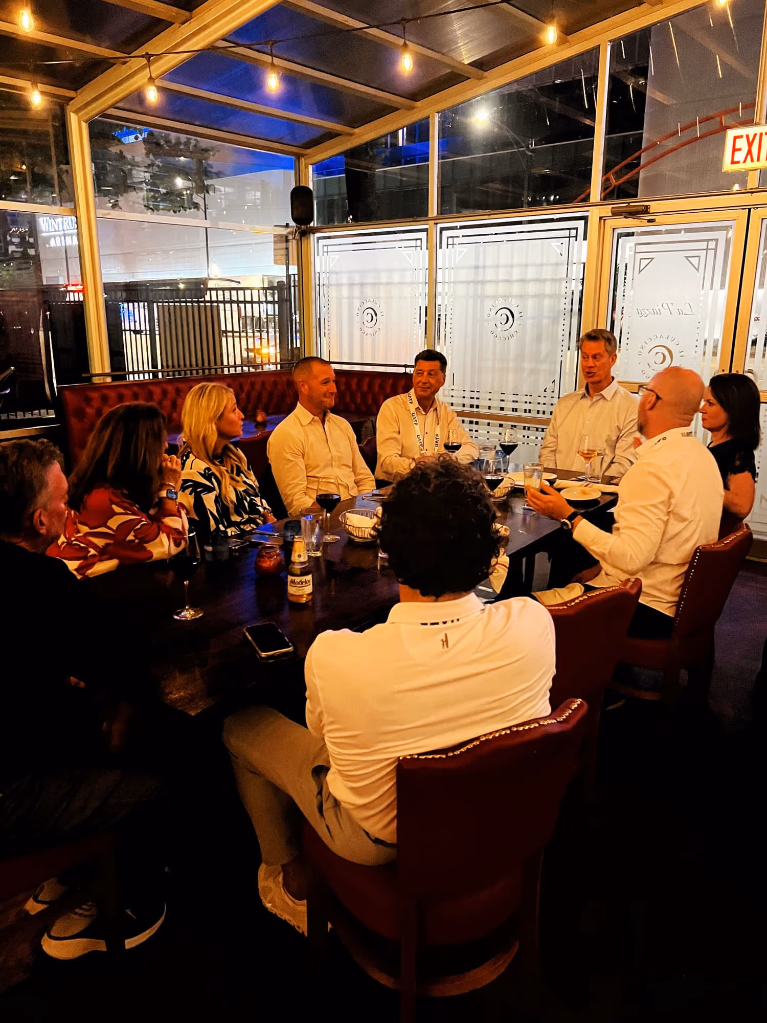Group of eight people sitting around a dark wooden table at a restaurant with drinks and conversation.