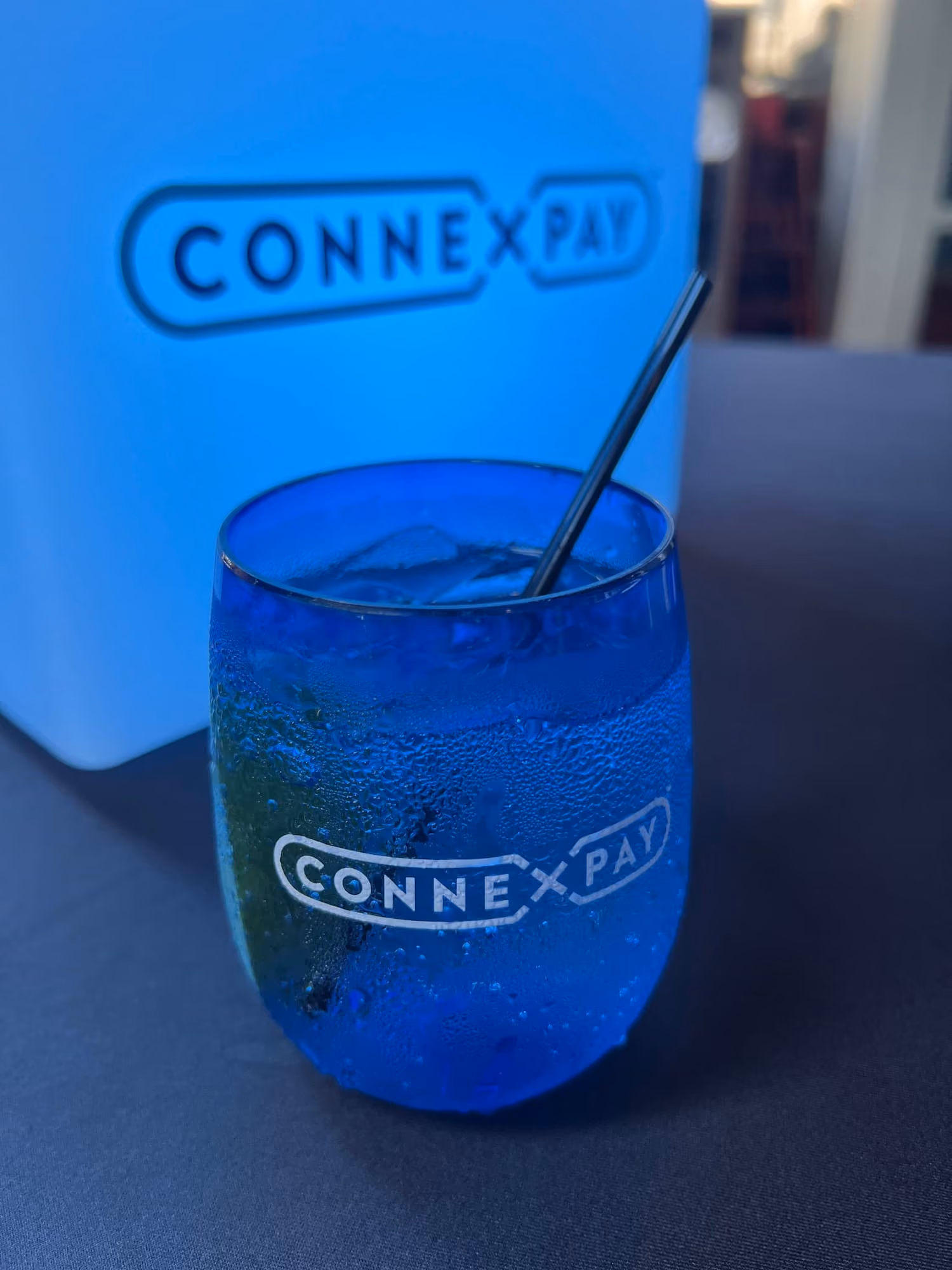 Condensation-covered blue CONNEX PAY glass with a metal straw on a dark surface with blurred background.