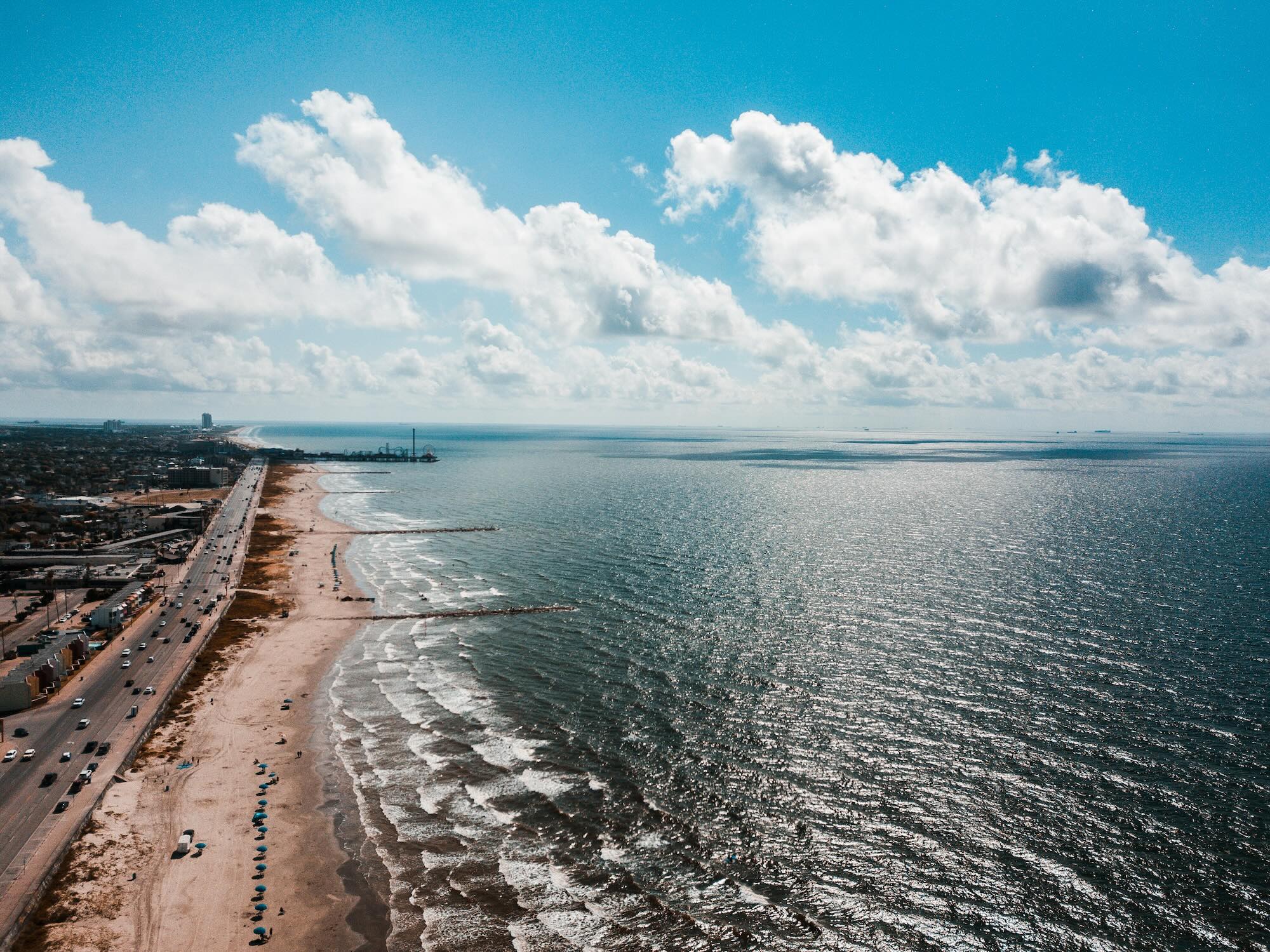 Aerial view of Galveston’s Seawall Boulevard and sandy beach with rows of umbrellas, gentle Gulf waves, and an amusement pier in the distance under a bright, cloud-filled sky.