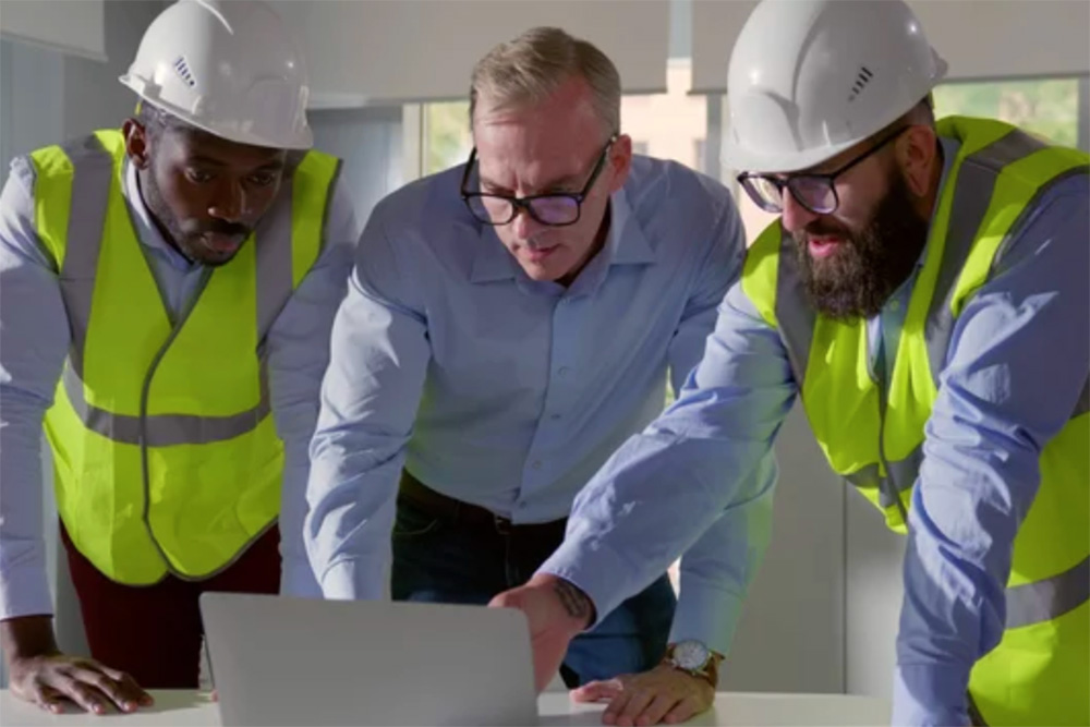 Three construction professionals—two in hard hats and high-visibility vests—lean over a table, reviewing plans on a laptop in a bright office.