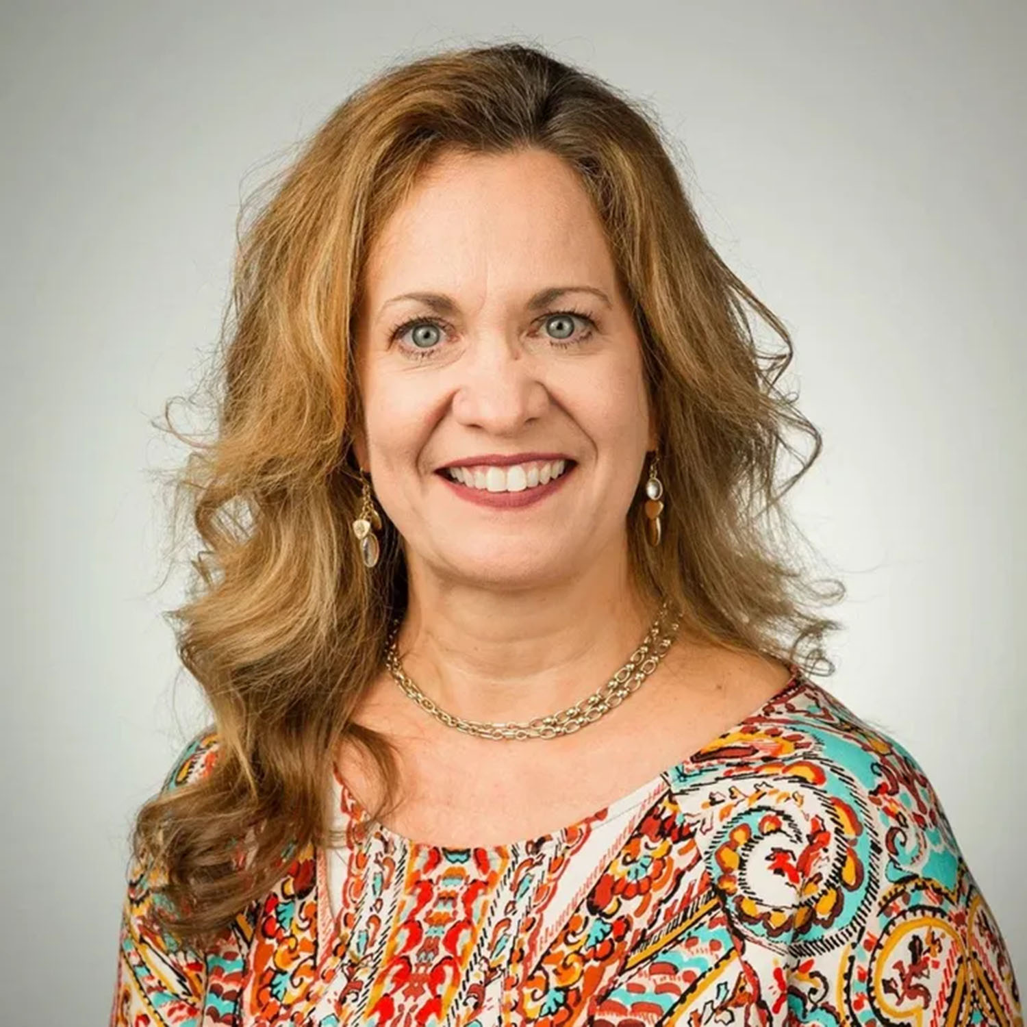 Smiling woman with wavy light-brown hair wearing gold earrings, a chain necklace, and a colorful patterned blouse, posed against a plain light gray background.
