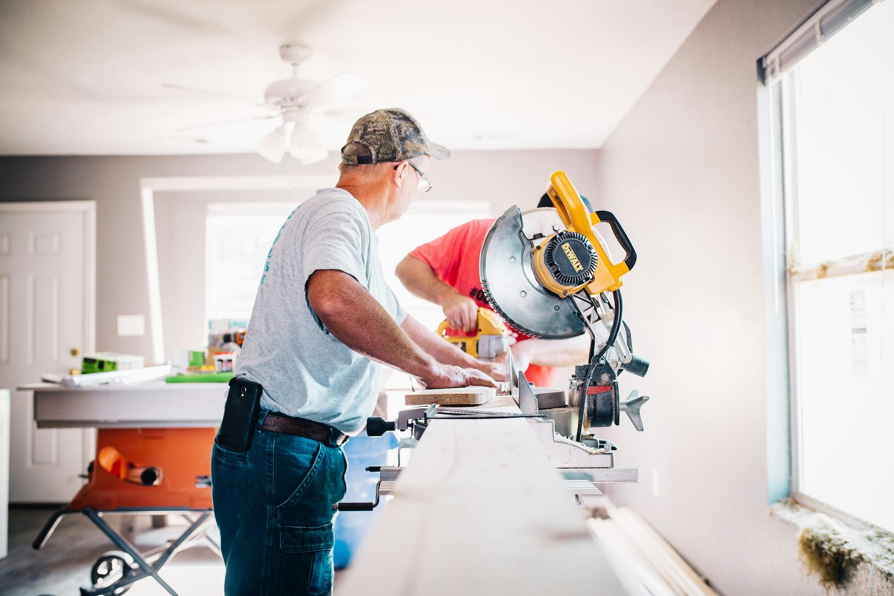 Two construction workers cut a board on a miter saw in a bright, partially renovated room with tools and lumber on the workbench.
