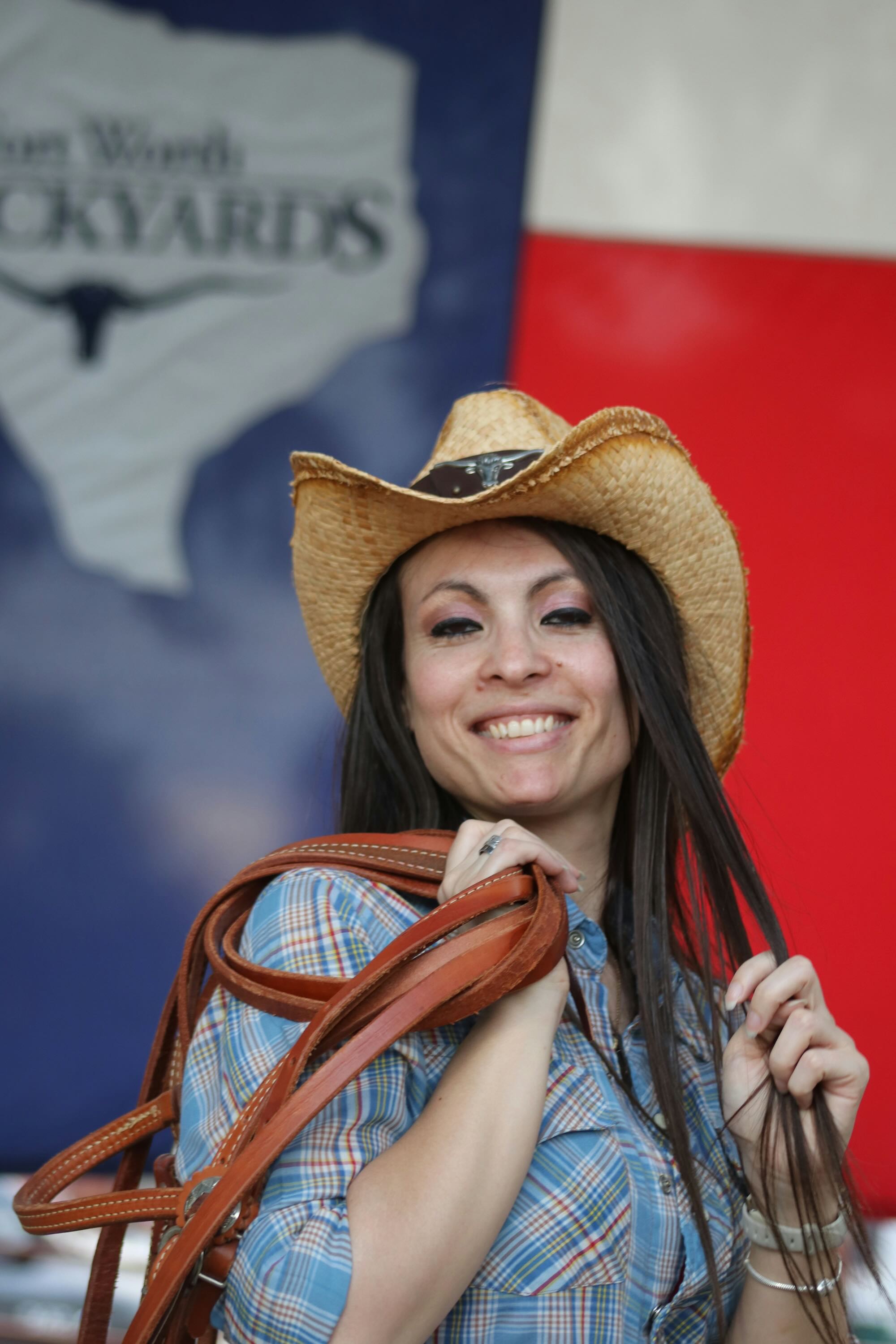 Smiling woman in a straw cowboy hat and plaid shirt holding leather reins over her shoulder, standing before a Texas flag and Fort Worth Stockyards sign.
