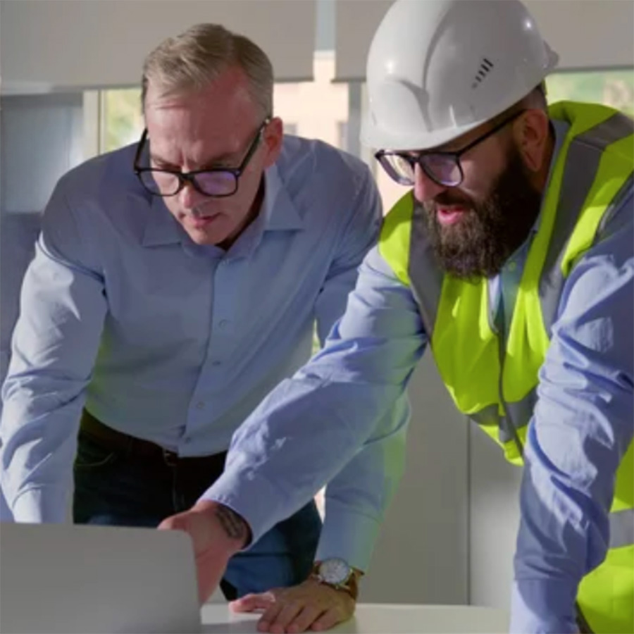 Two construction professionals lean over a laptop, one in a hard hat and safety vest and the other in a blue dress shirt and glasses, reviewing project plans in a bright office.