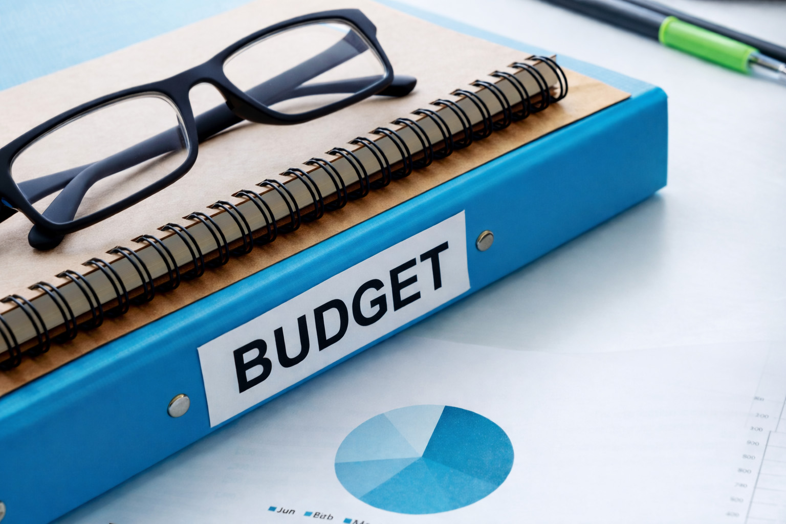 Blue binder labeled "Budget" with reading glasses on top, resting on financial reports