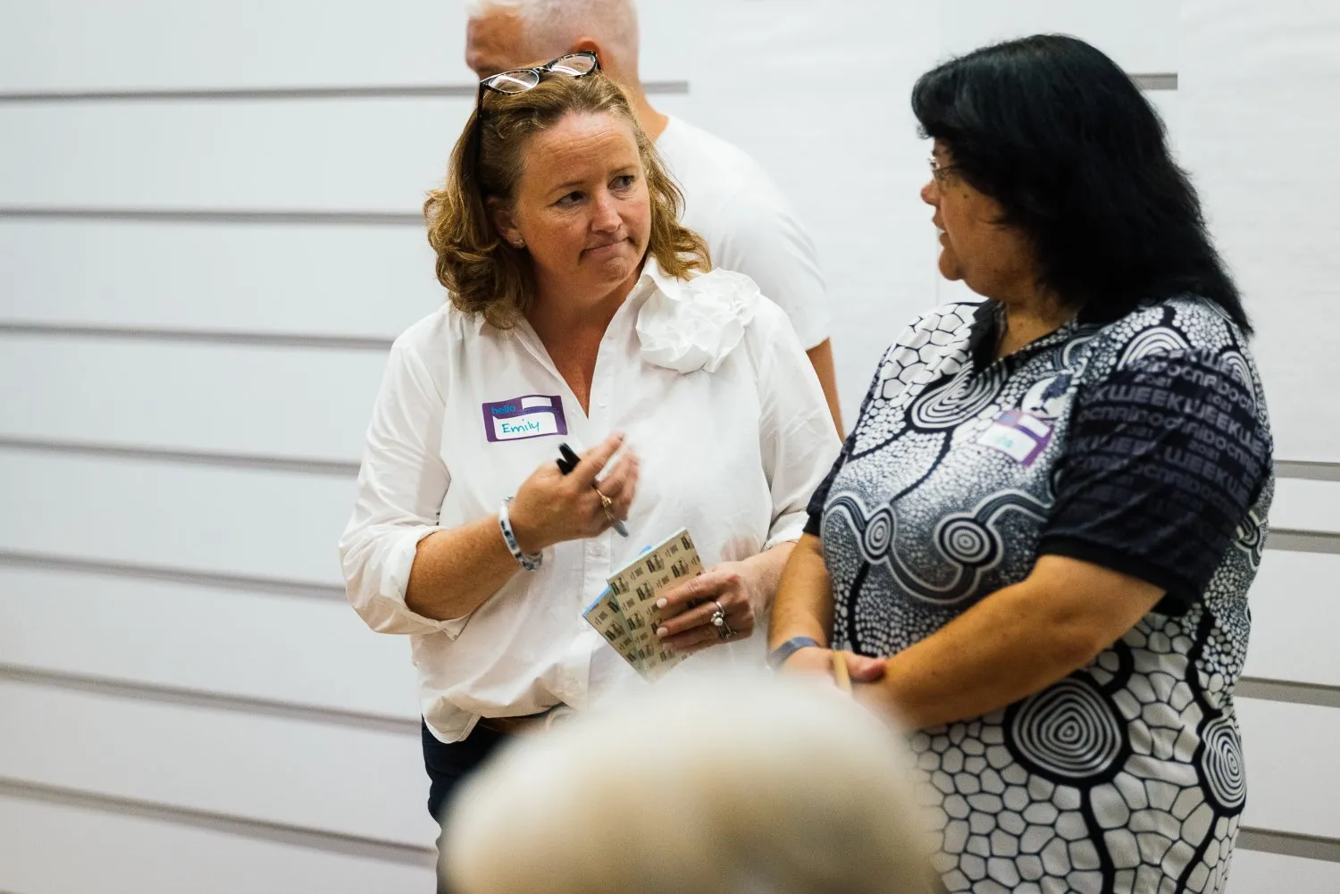 Two women engaged in conversation in a bright indoor setting, one holding a notebook and wearing a white blouse with a name tag labeled Emily.