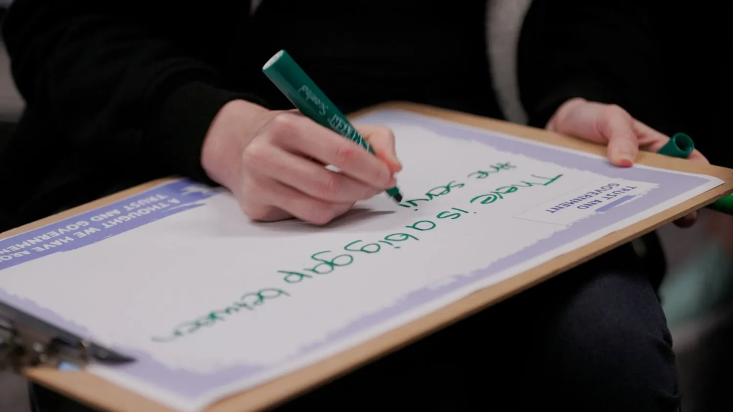 Person writing ‘There is a big gap between the sort’ on a clipboard with a green marker.