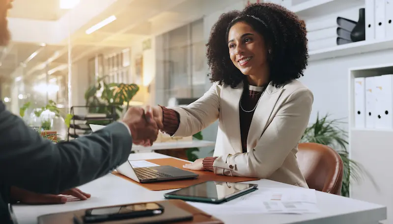 Smiling woman in a white blazer shaking hands with a man across a desk in a bright office.
