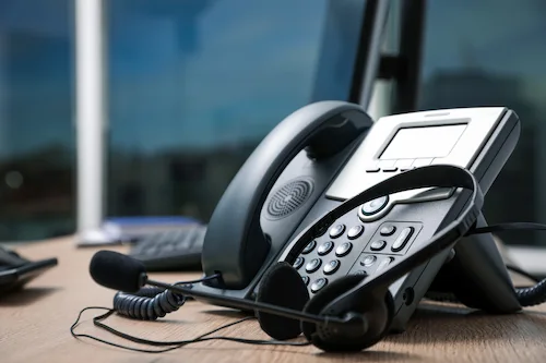 Desk phone with a headset resting on a wooden table in an office setting.