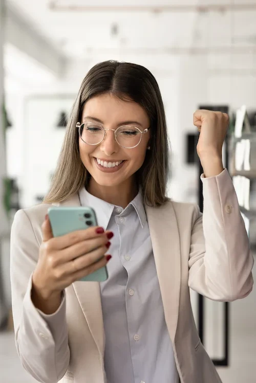 Smiling businesswoman wearing glasses and blazer excitedly looking at her smartphone and raising a fist in success.