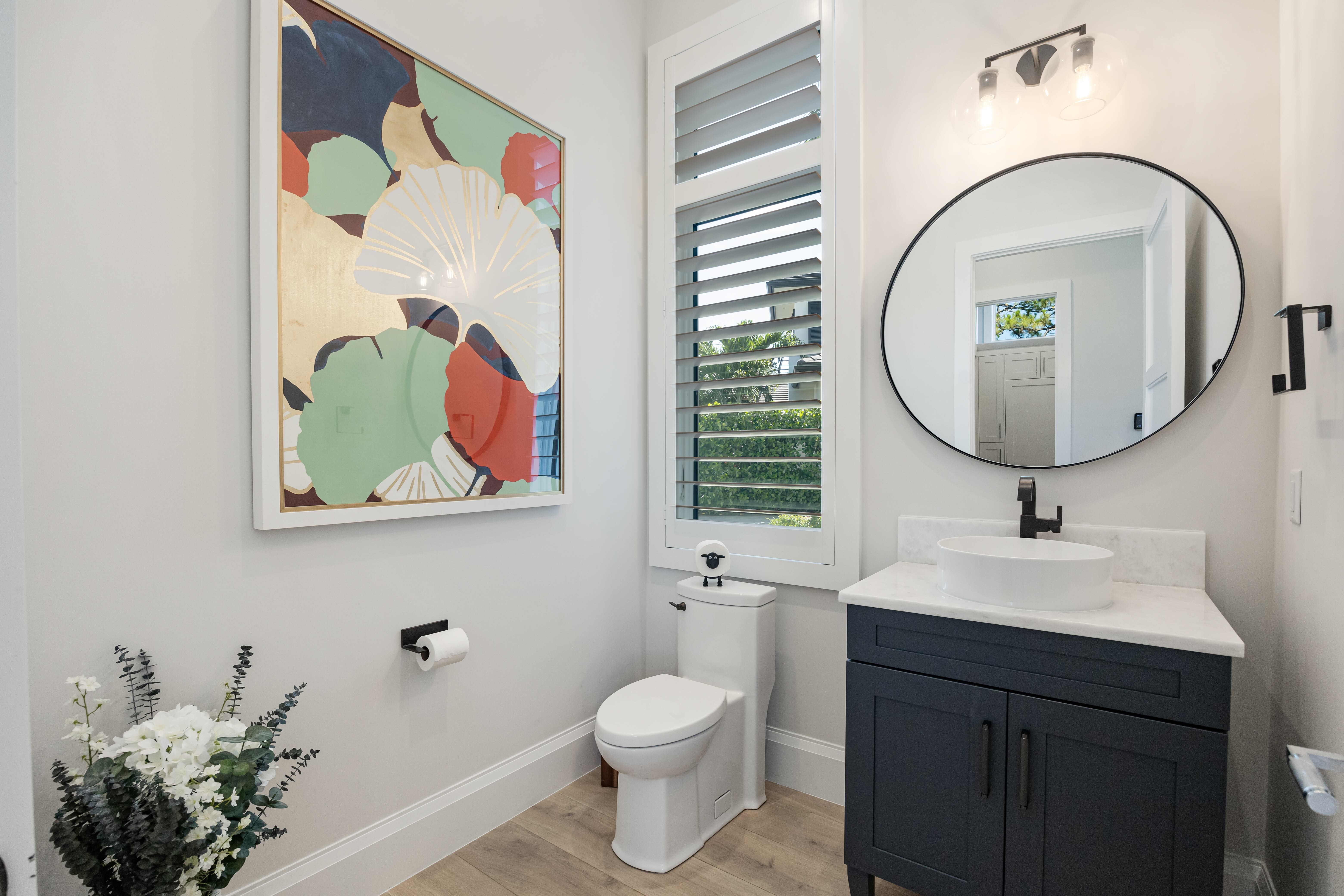 Modern bathroom with white toilet, navy blue vanity, round mirror, window with blinds, colorful abstract wall art, and a plant with white flowers.