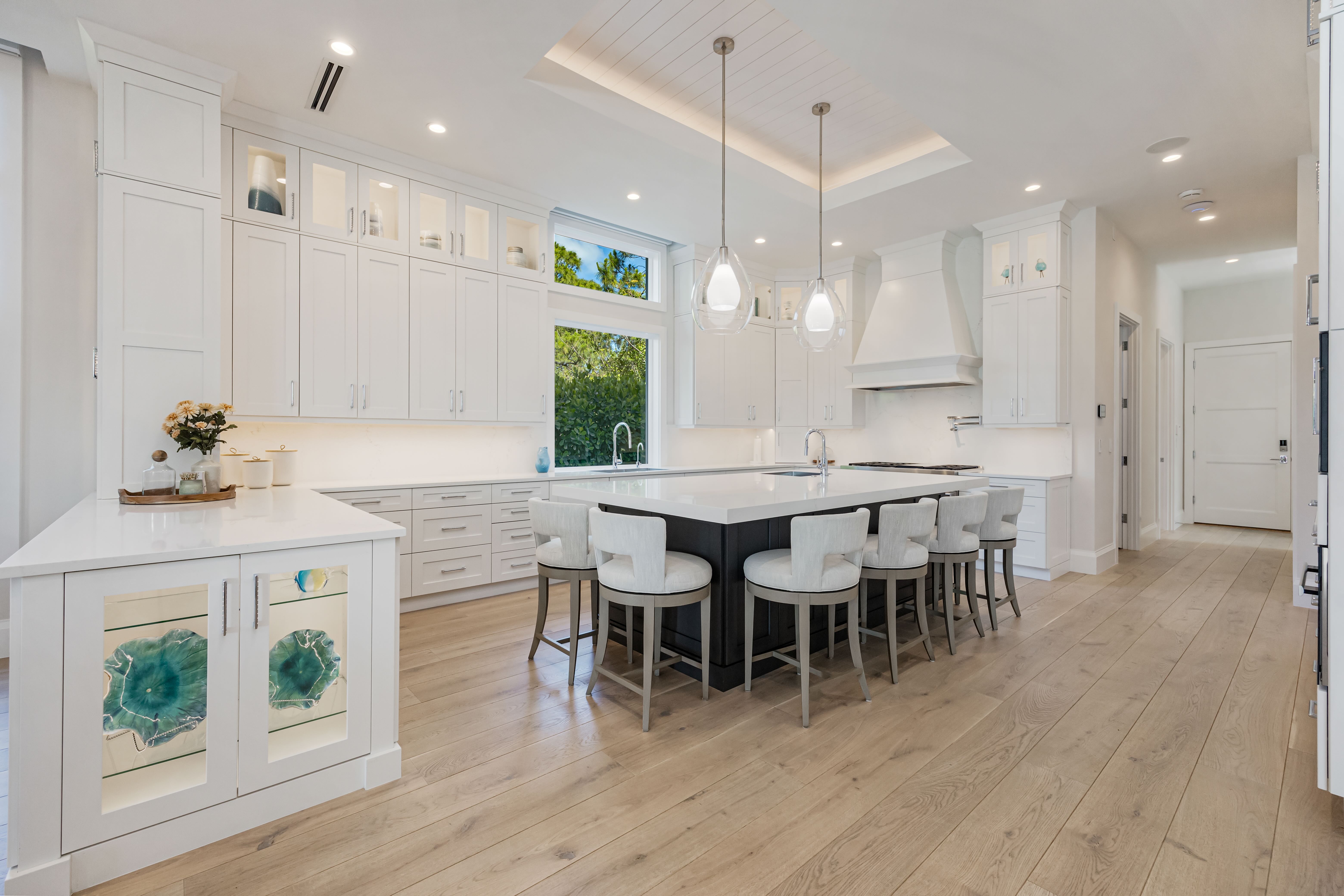 Bright modern kitchen with large white island, six cushioned stools, white cabinetry, light wood flooring, and pendant lights.
