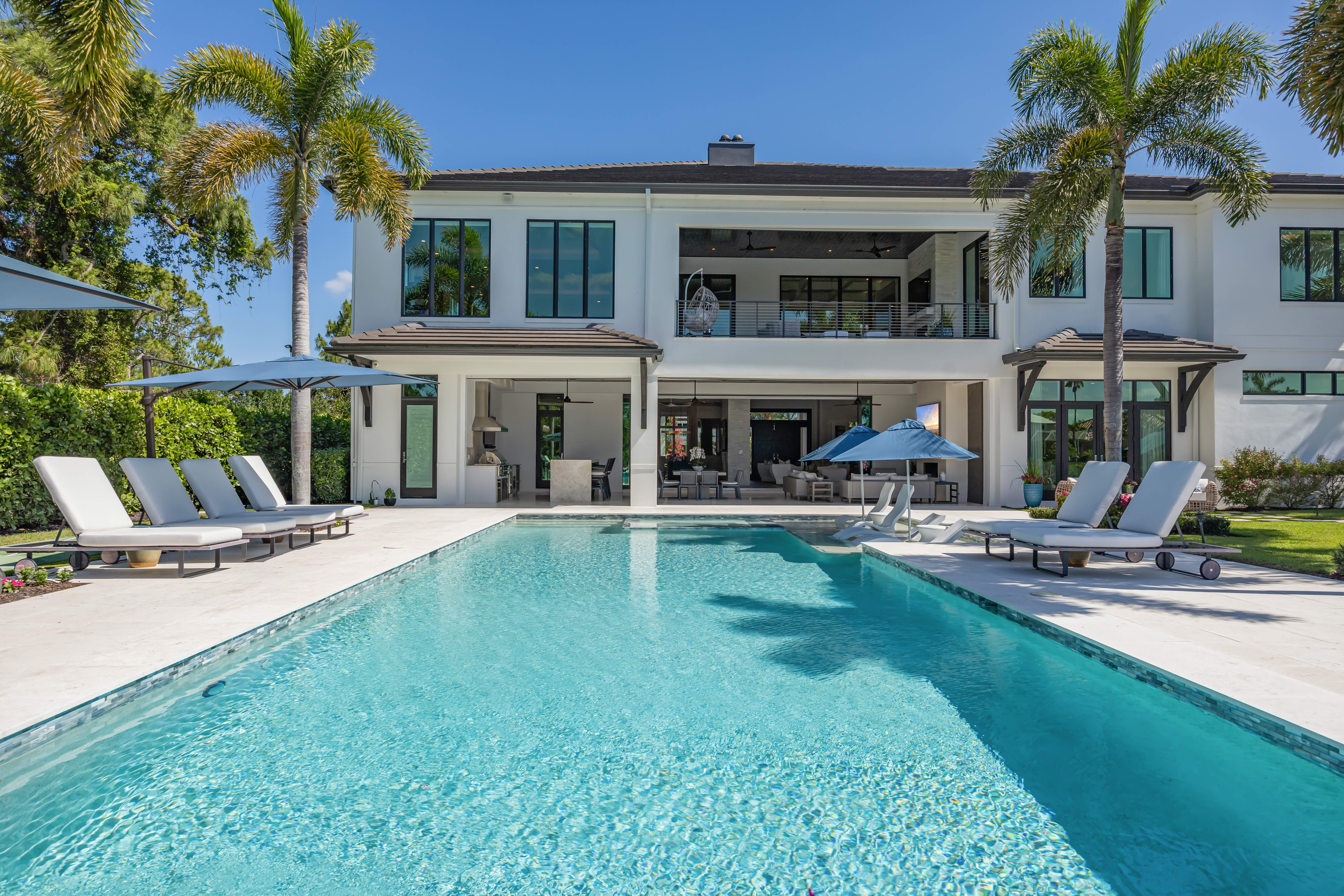 Modern two-story house with a large swimming pool, lounge chairs, umbrellas, and palm trees in the backyard on a sunny day.