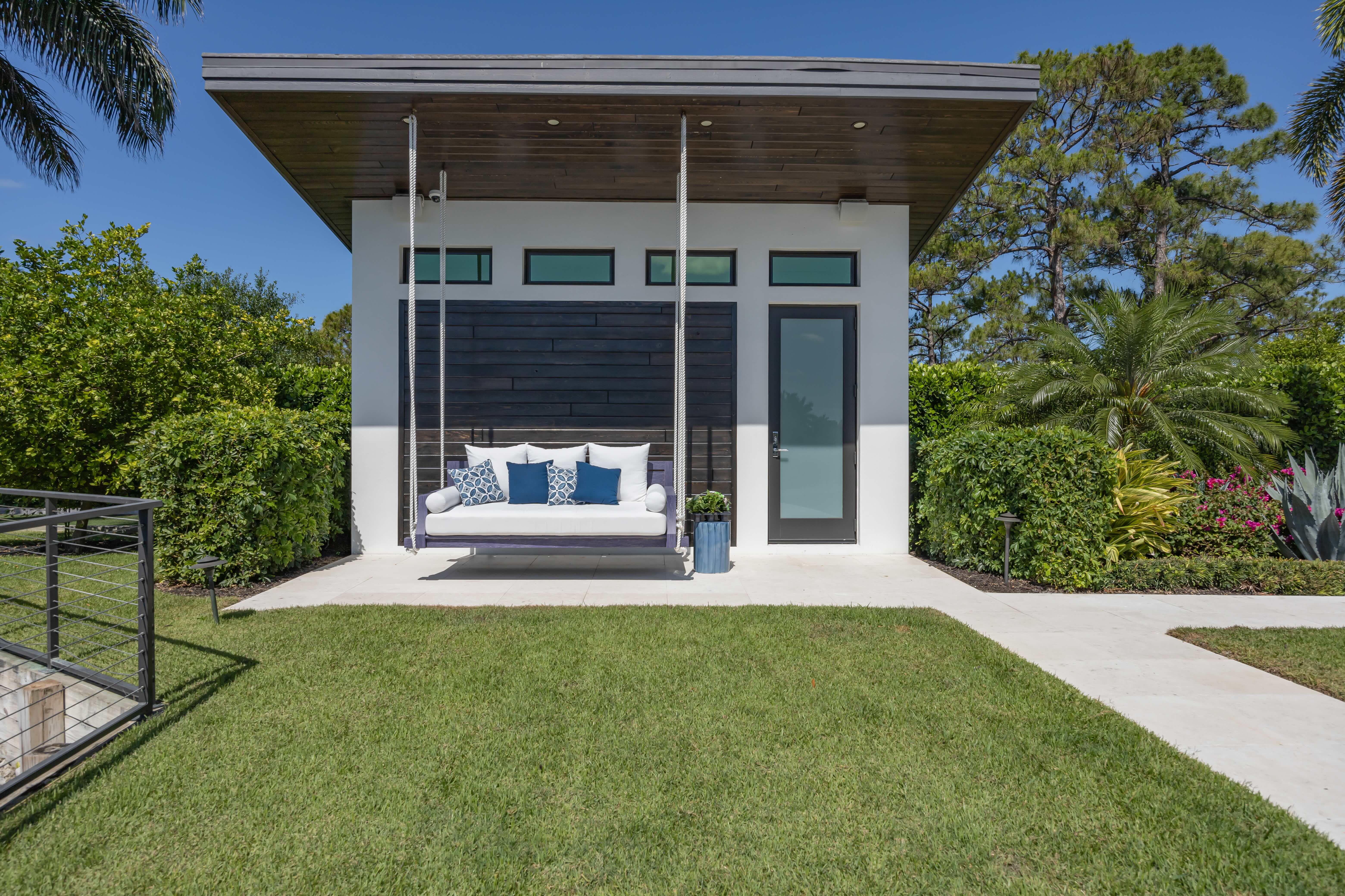 Modern white pool house with a hanging outdoor sofa accented by blue and white pillows beside a frosted glass door, surrounded by green shrubs and trees under a clear blue sky.